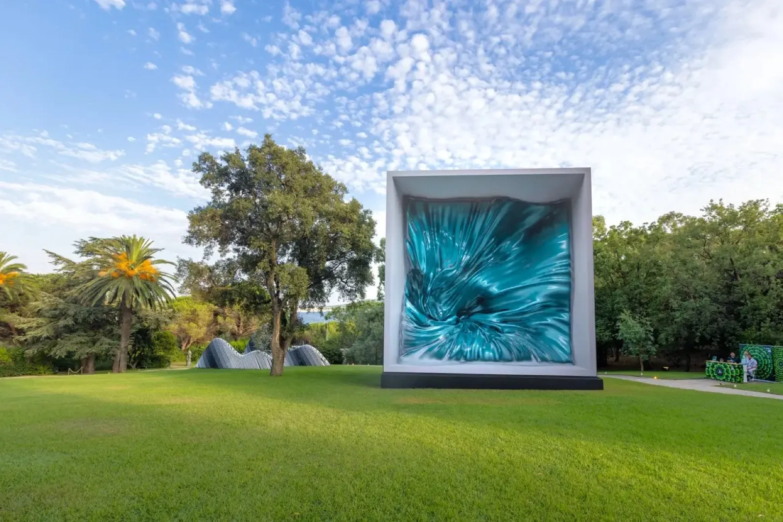 Large white-framed LED cube showing swirling teal fluid digital art installed on a park lawn