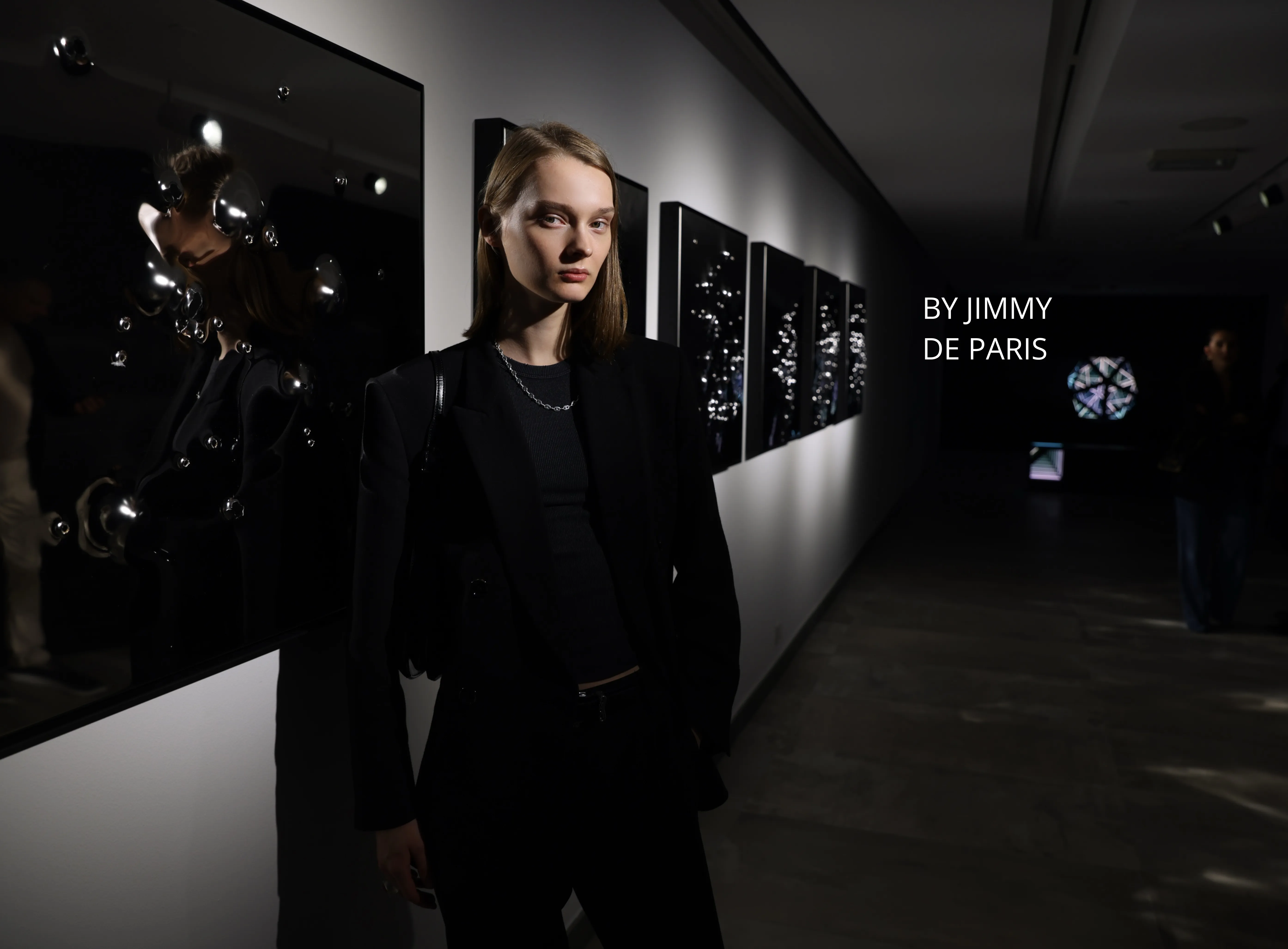 Woman in black in a dark gallery corridor, 'by Jimmy de Paris' wall text