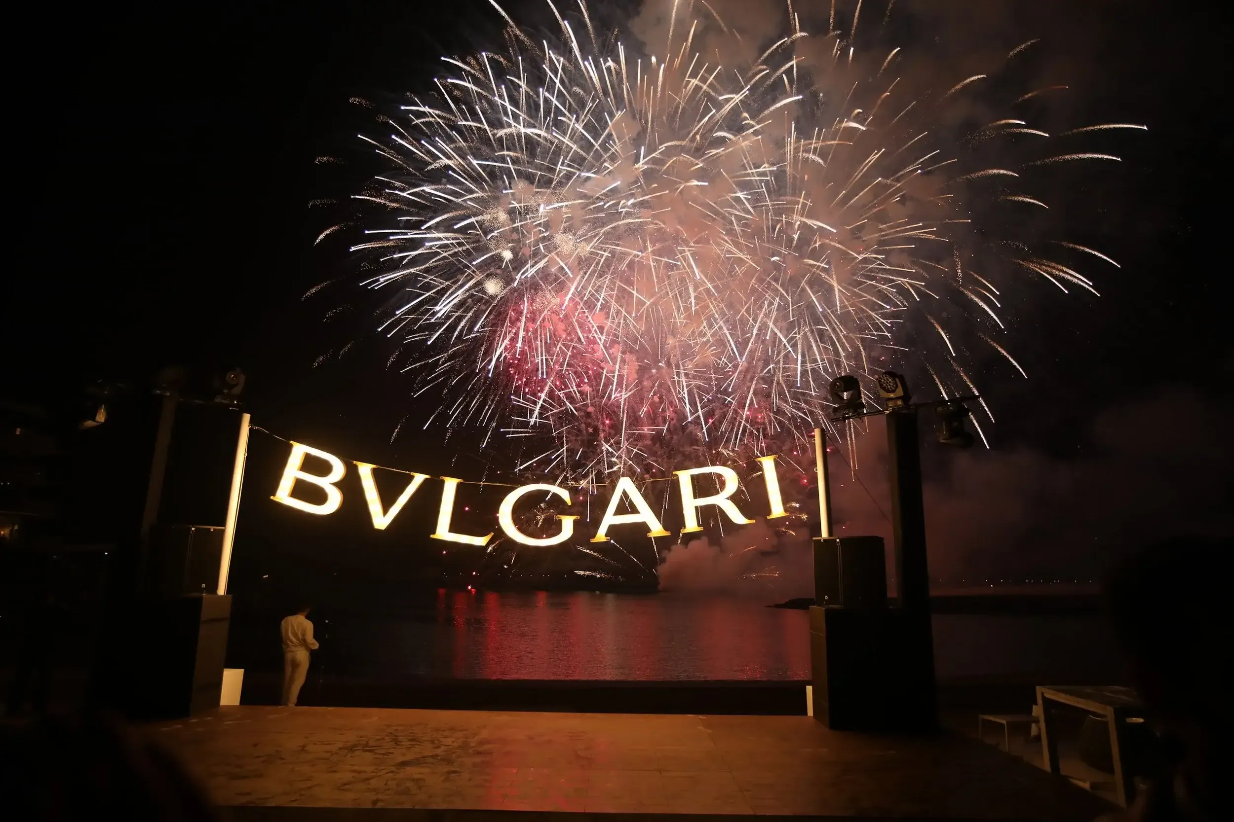 Illuminated BVLGARI letters on a waterfront stage with fireworks exploding over the water at night