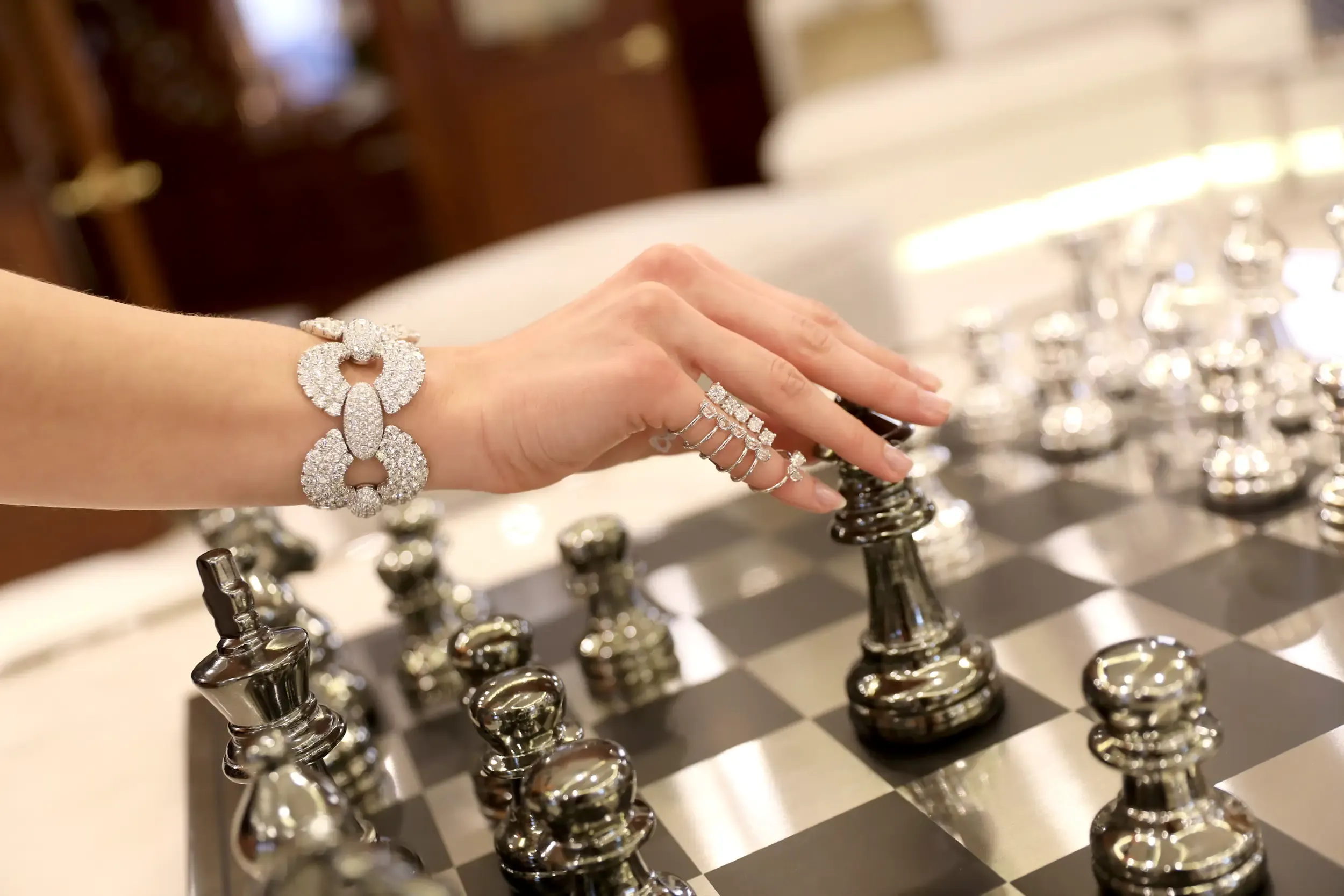 Detail of a diamond bracelet and rings as a hand reaches for a king on a metallic chessboard at a luxury setting