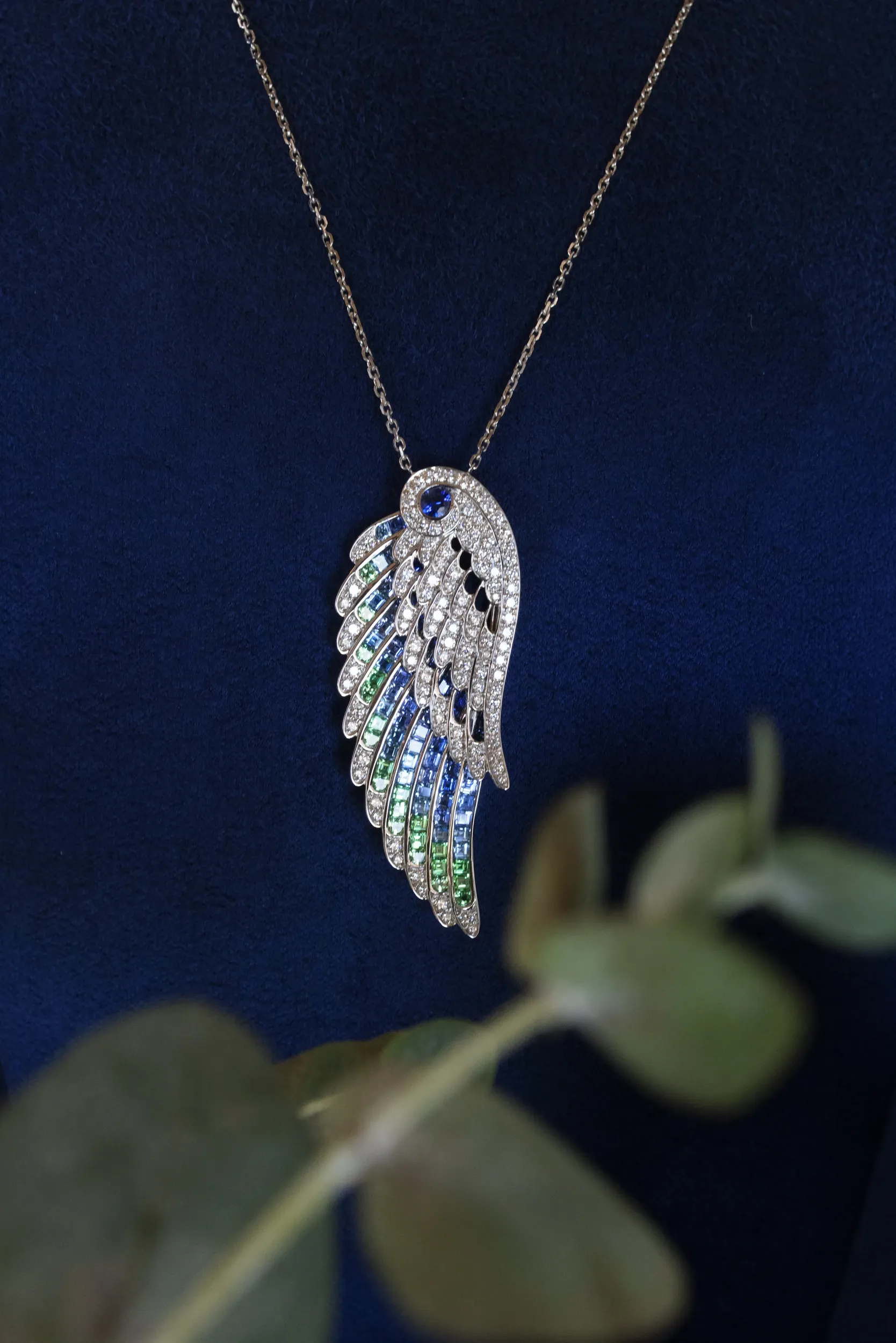 A feather-shaped jewelled pendant on a fine chain against dark blue fabric.