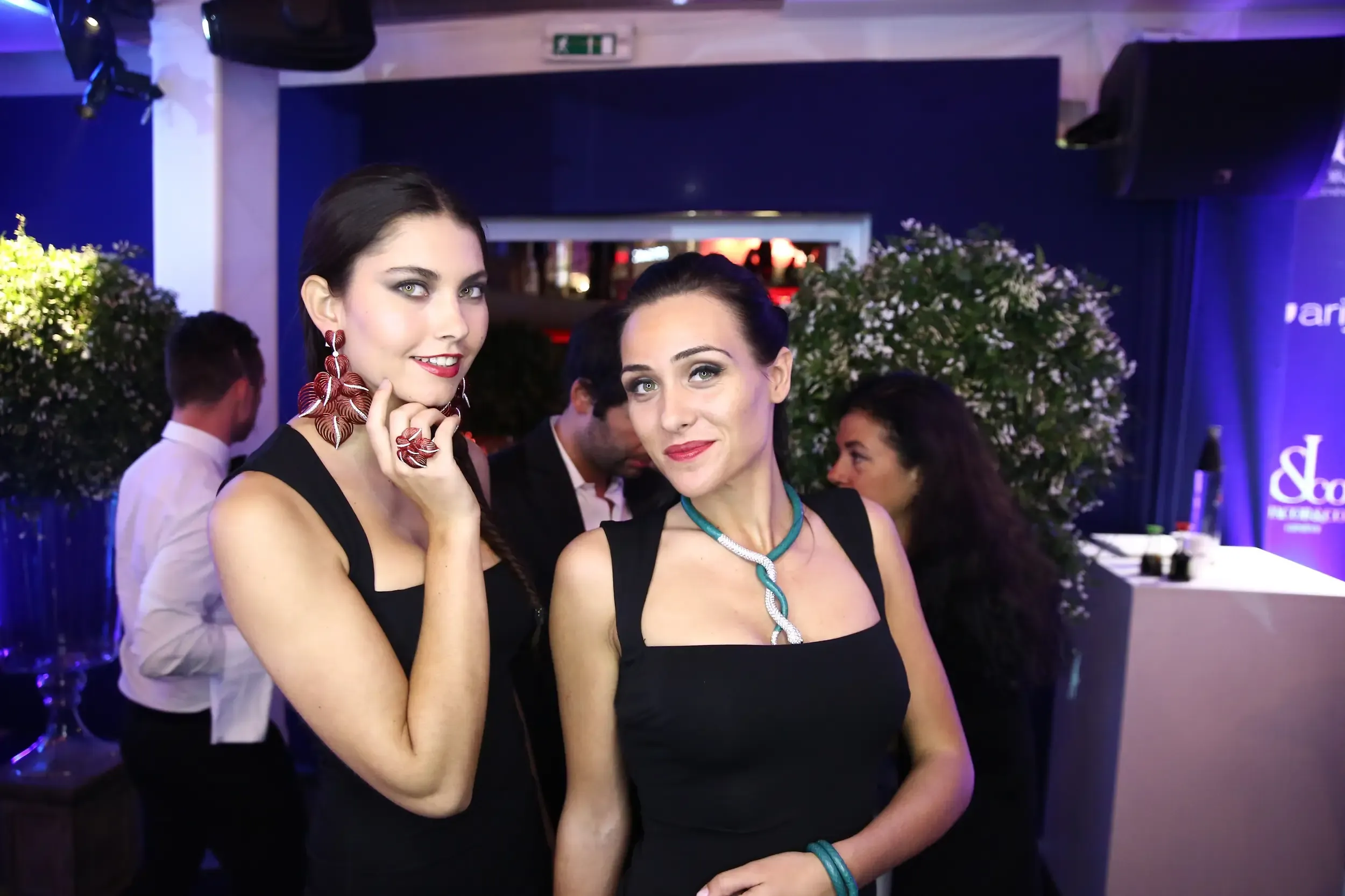 Two guests in black dresses wearing statement orchid earrings and a teal snake necklace at a Jacob & Co event