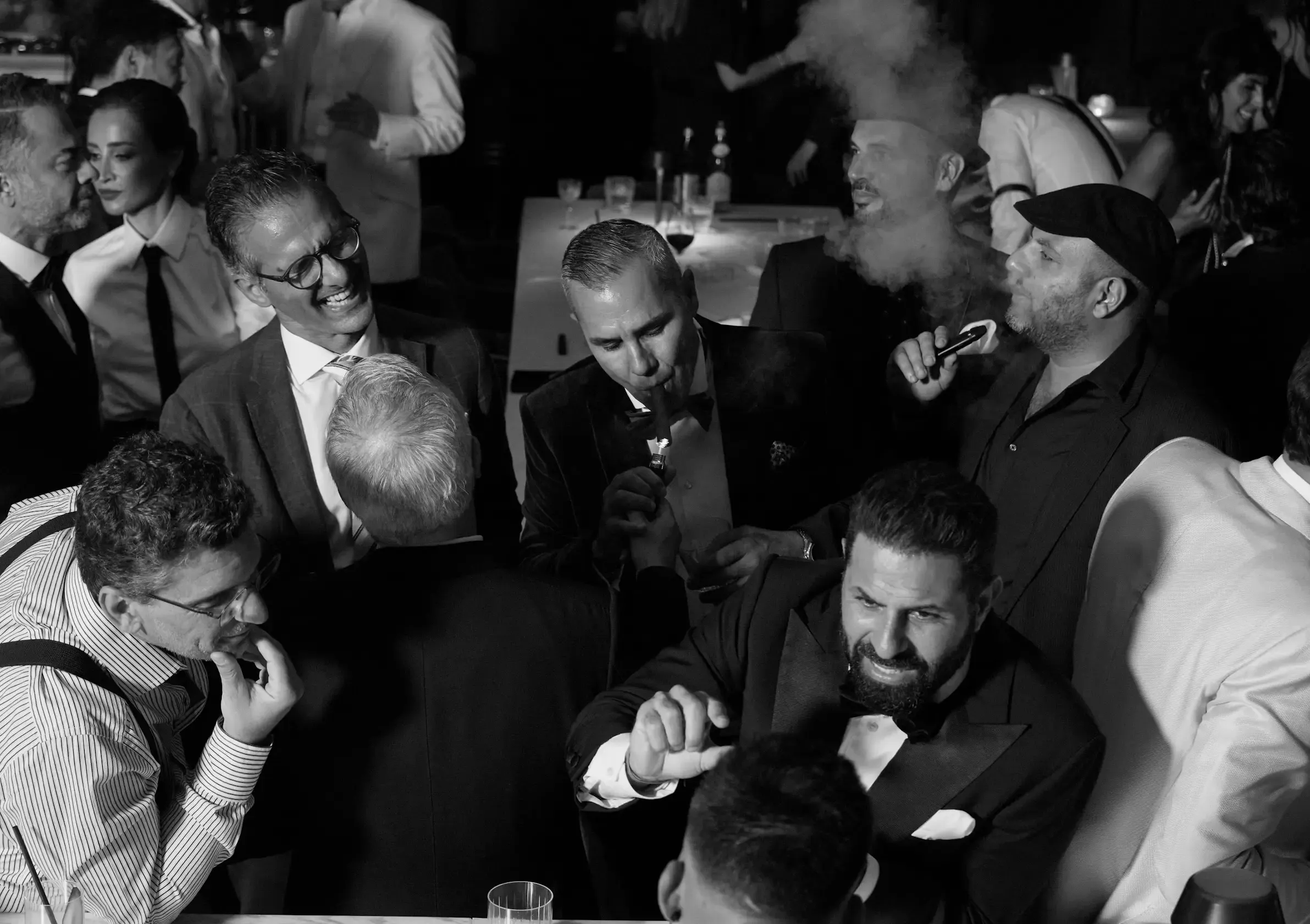 Black-and-white candid of tuxedoed guests lighting cigars and talking in a crowded night lounge