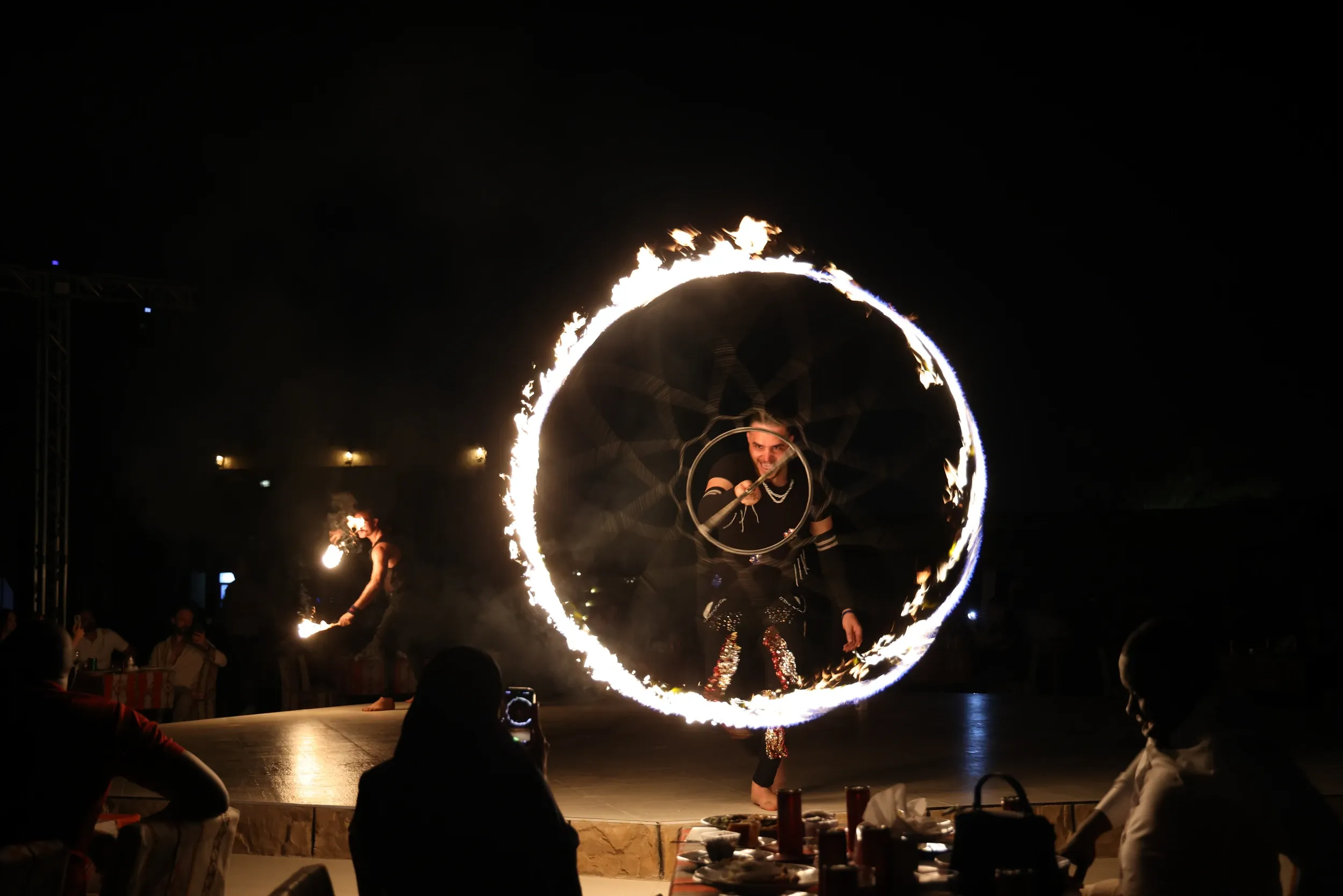 A fire performer framed by a circular trail of flame in front of seated guests.