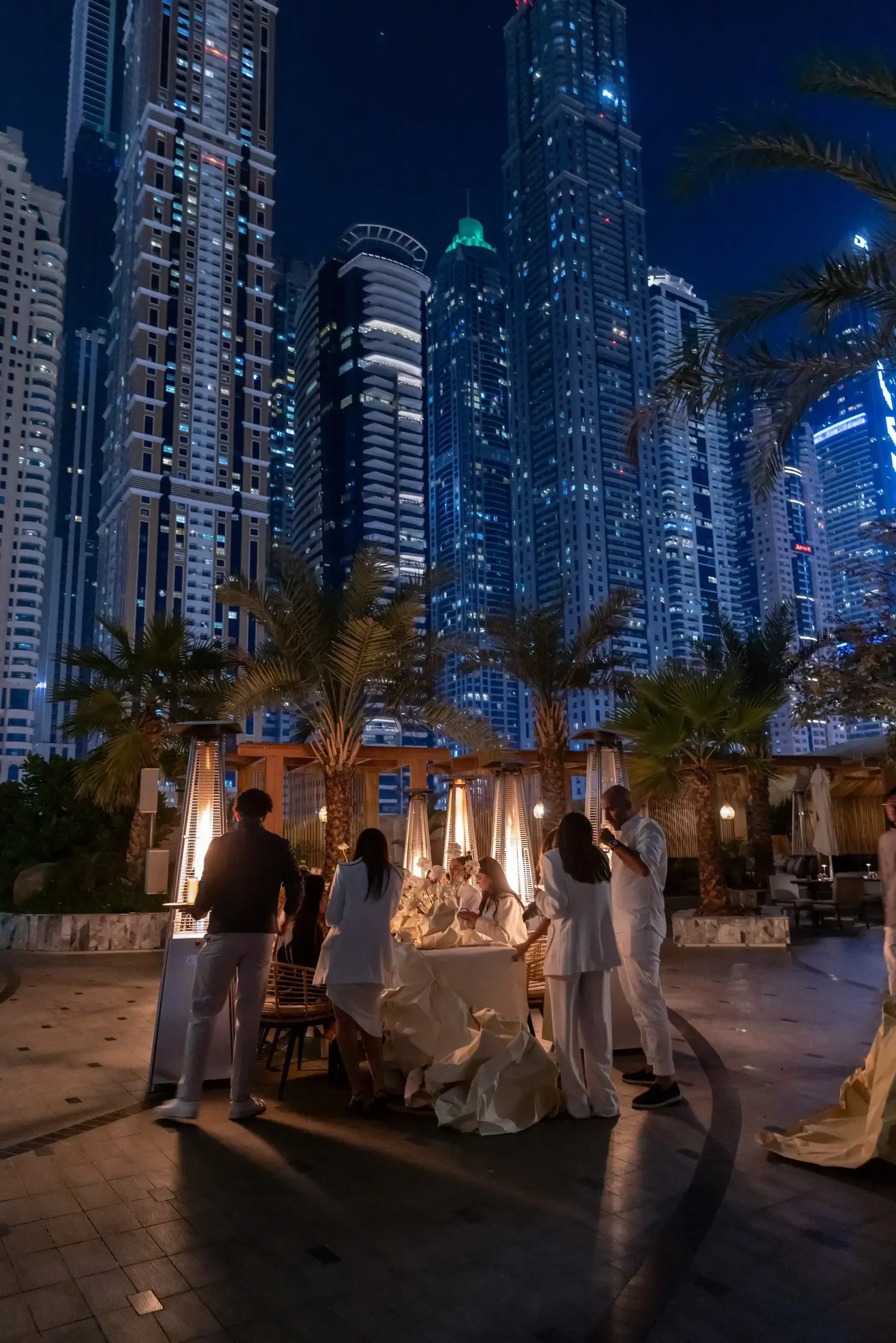 People standing around an outdoor dining table beneath illuminated towers.