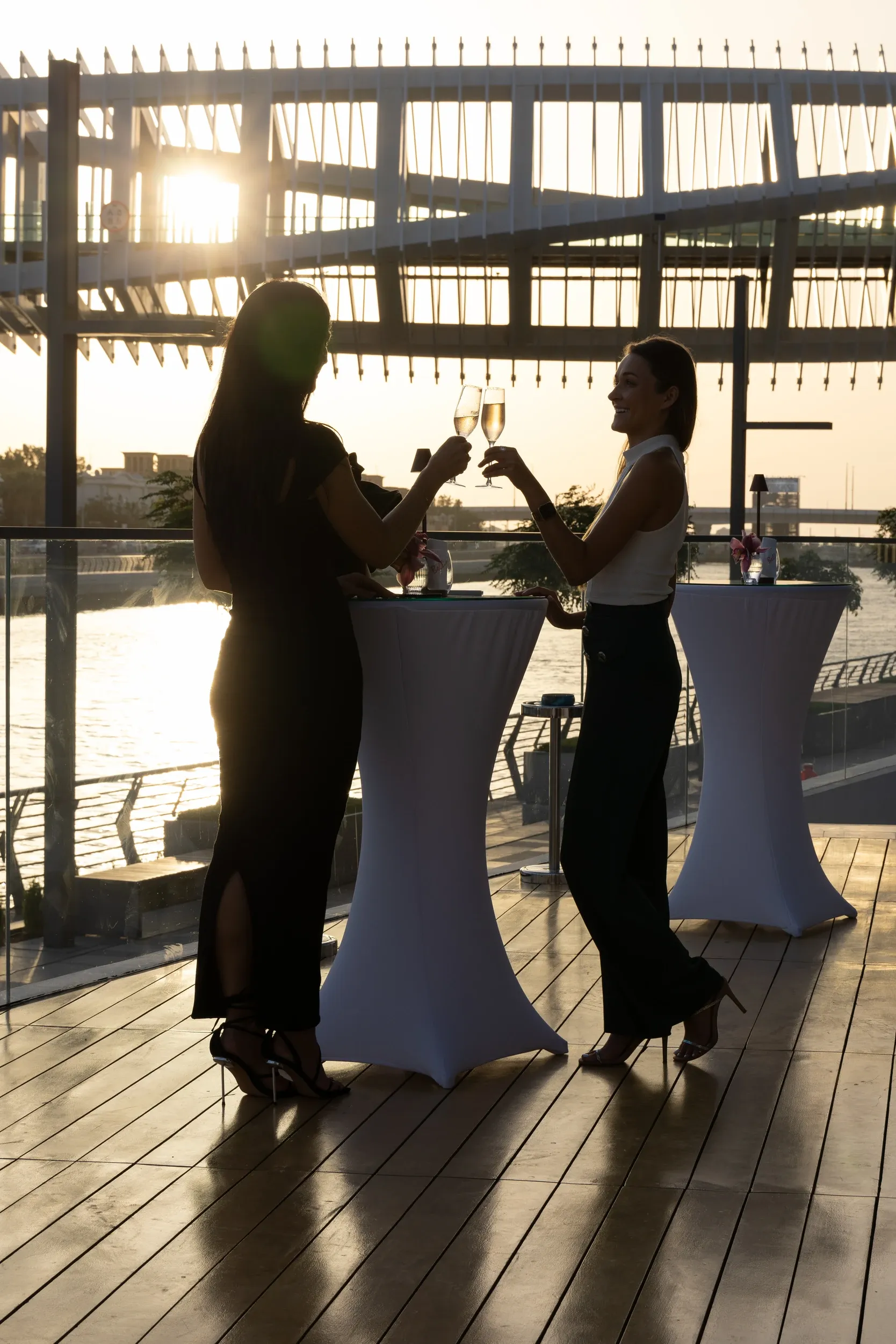 Two guests toasting on a waterfront terrace in low evening sunlight.