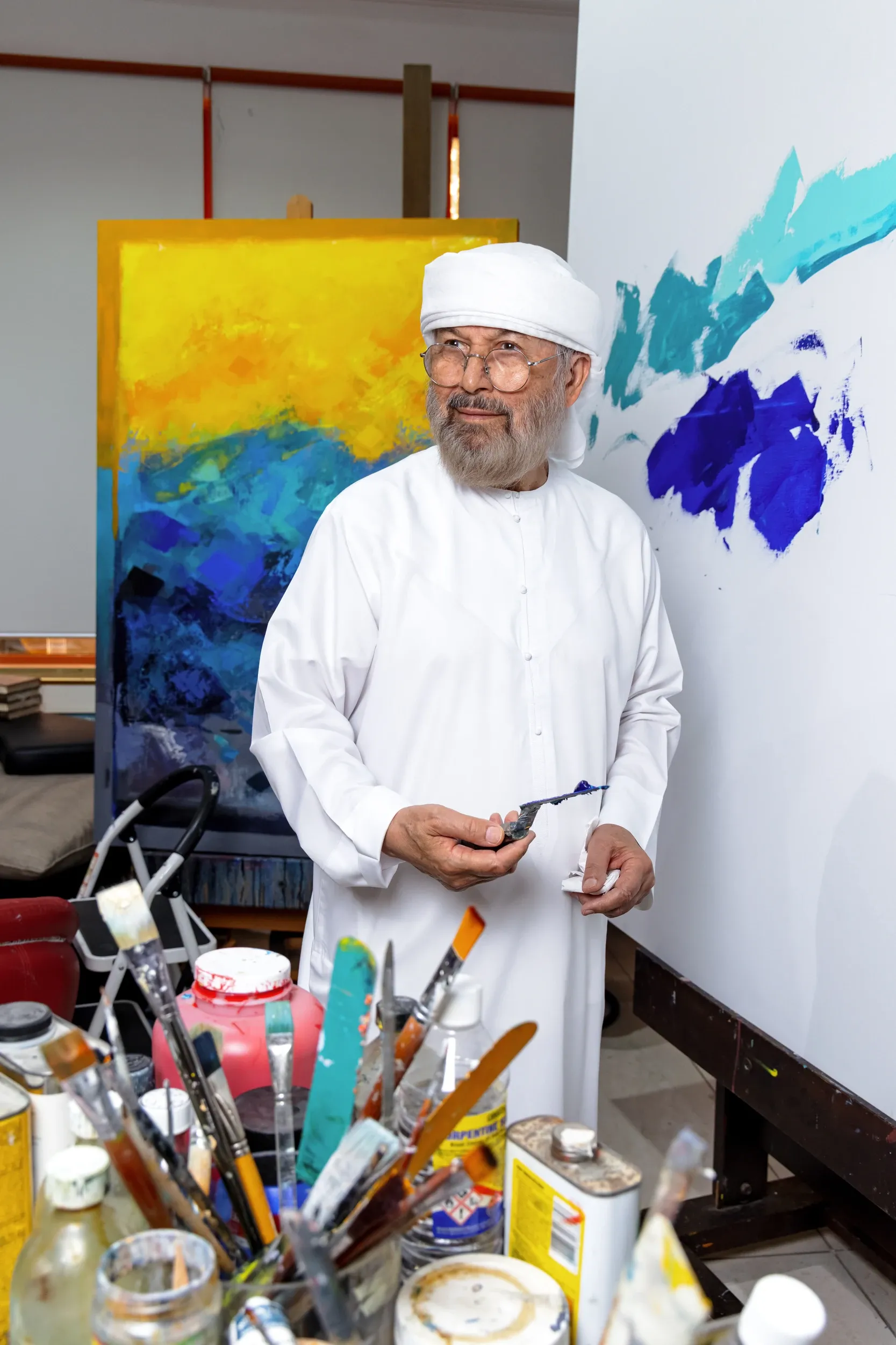 An artist in white holding a painting tool among brushes and colourful canvases.