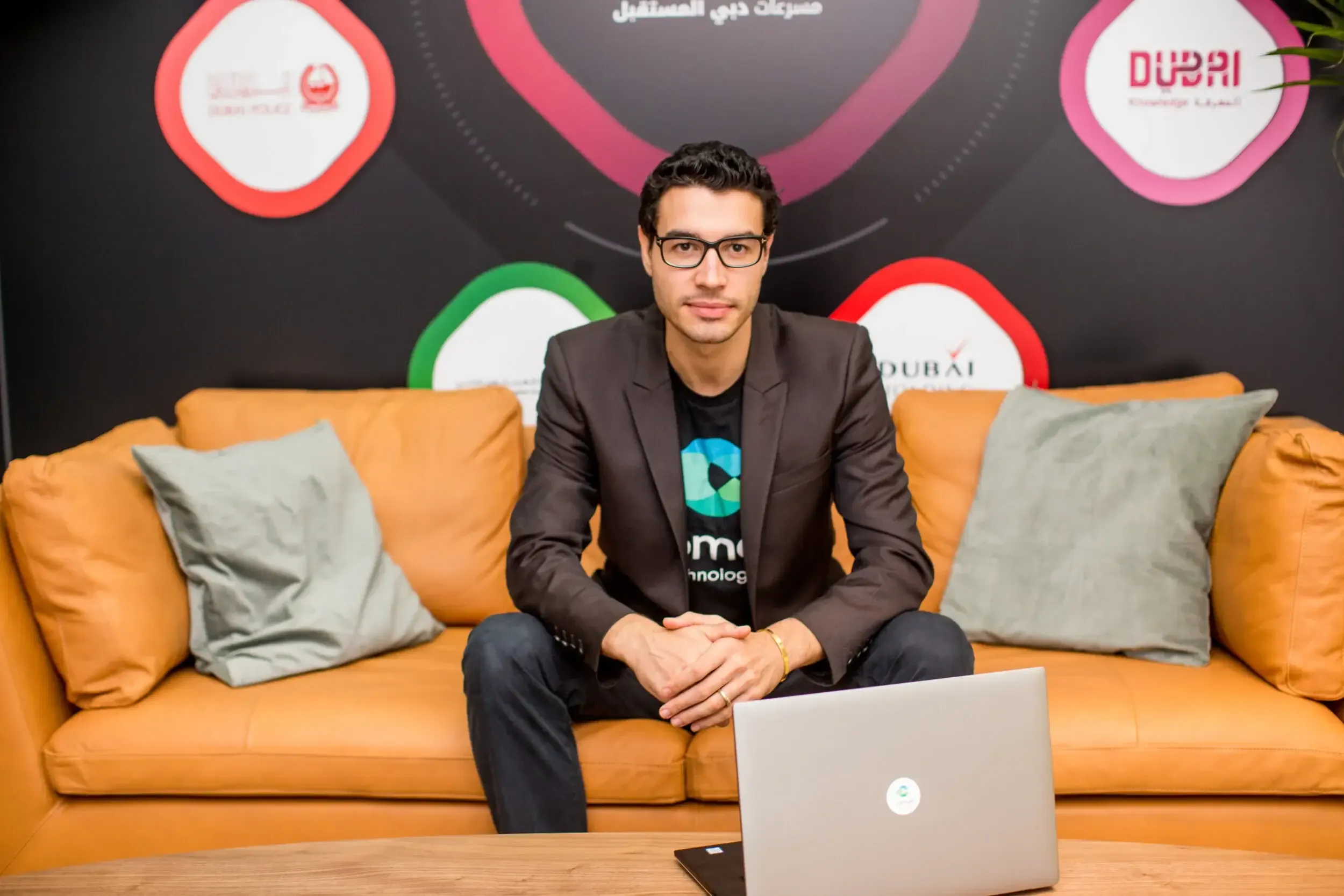 Tech founder in blazer and glasses on an orange sofa at Dubai Future Accelerators with laptop in foreground