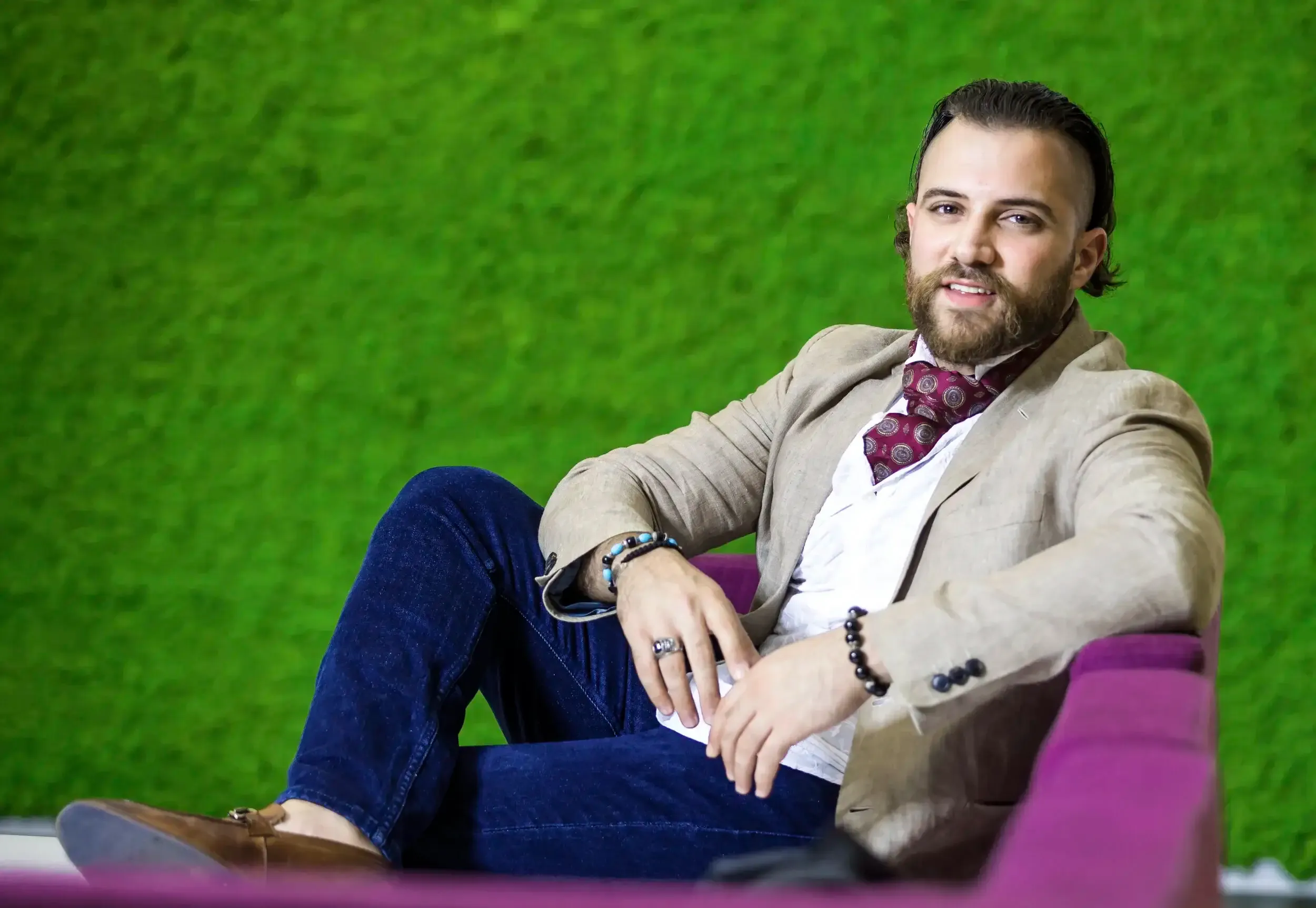 Relaxed executive portrait on a purple sofa against a textured green wall, linen blazer and burgundy ascot