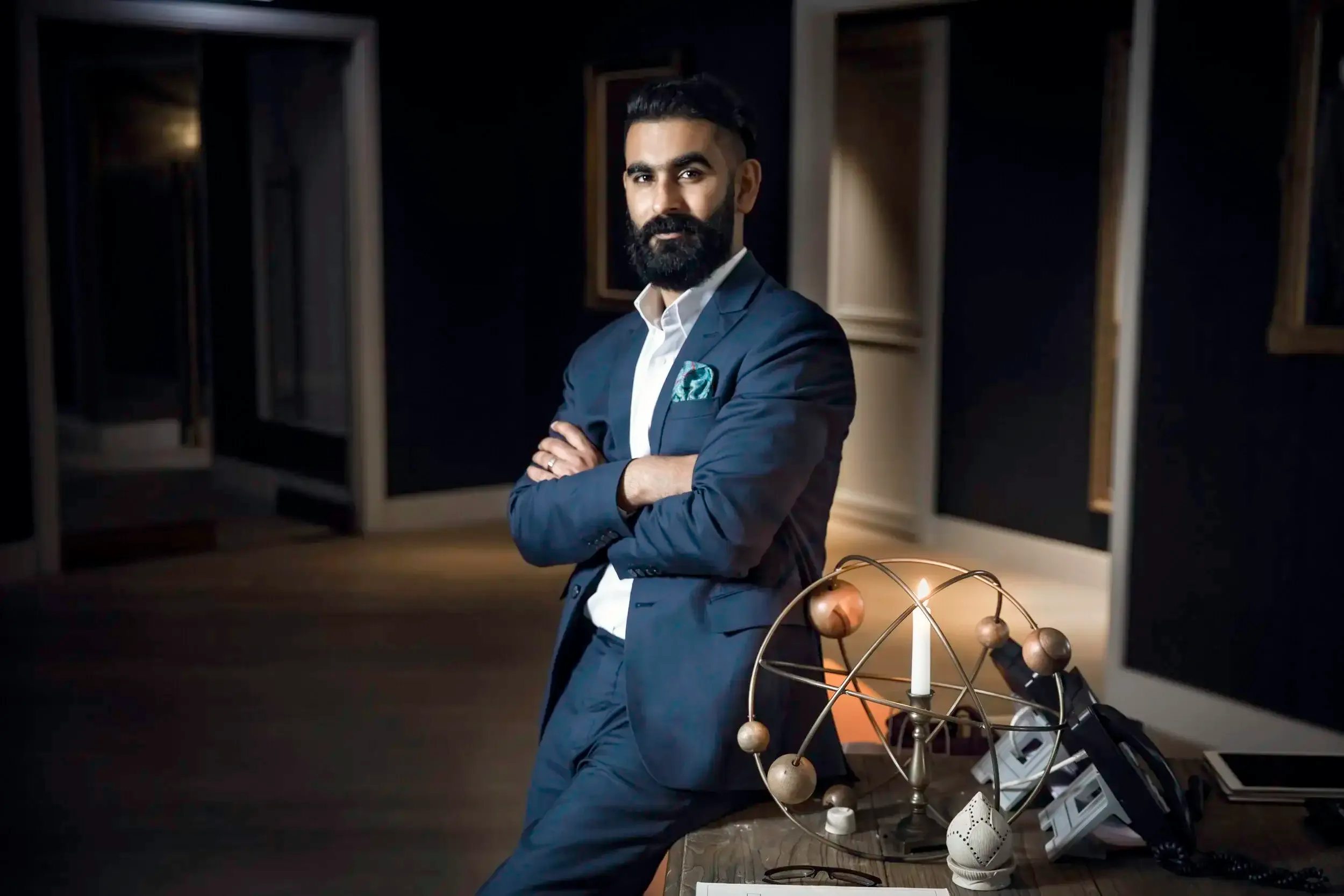 Moody executive portrait of a bearded man in a navy suit leaning on a candlelit wooden desk