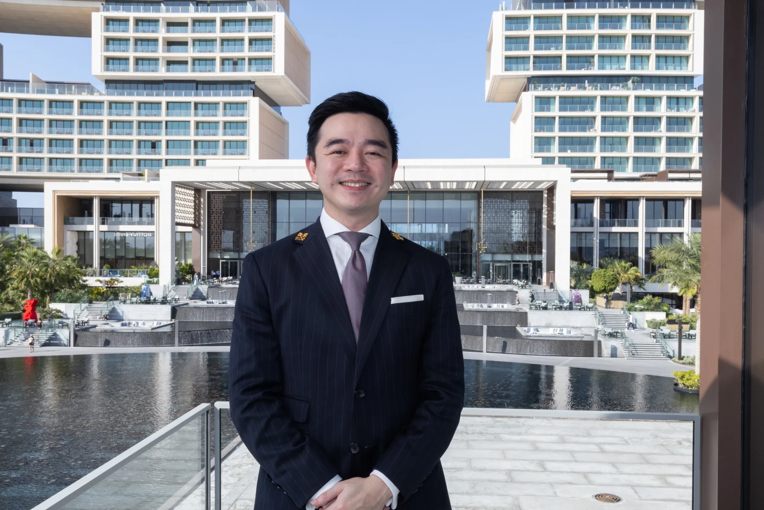 A man in a dark suit posed in front of a contemporary hotel and water feature.