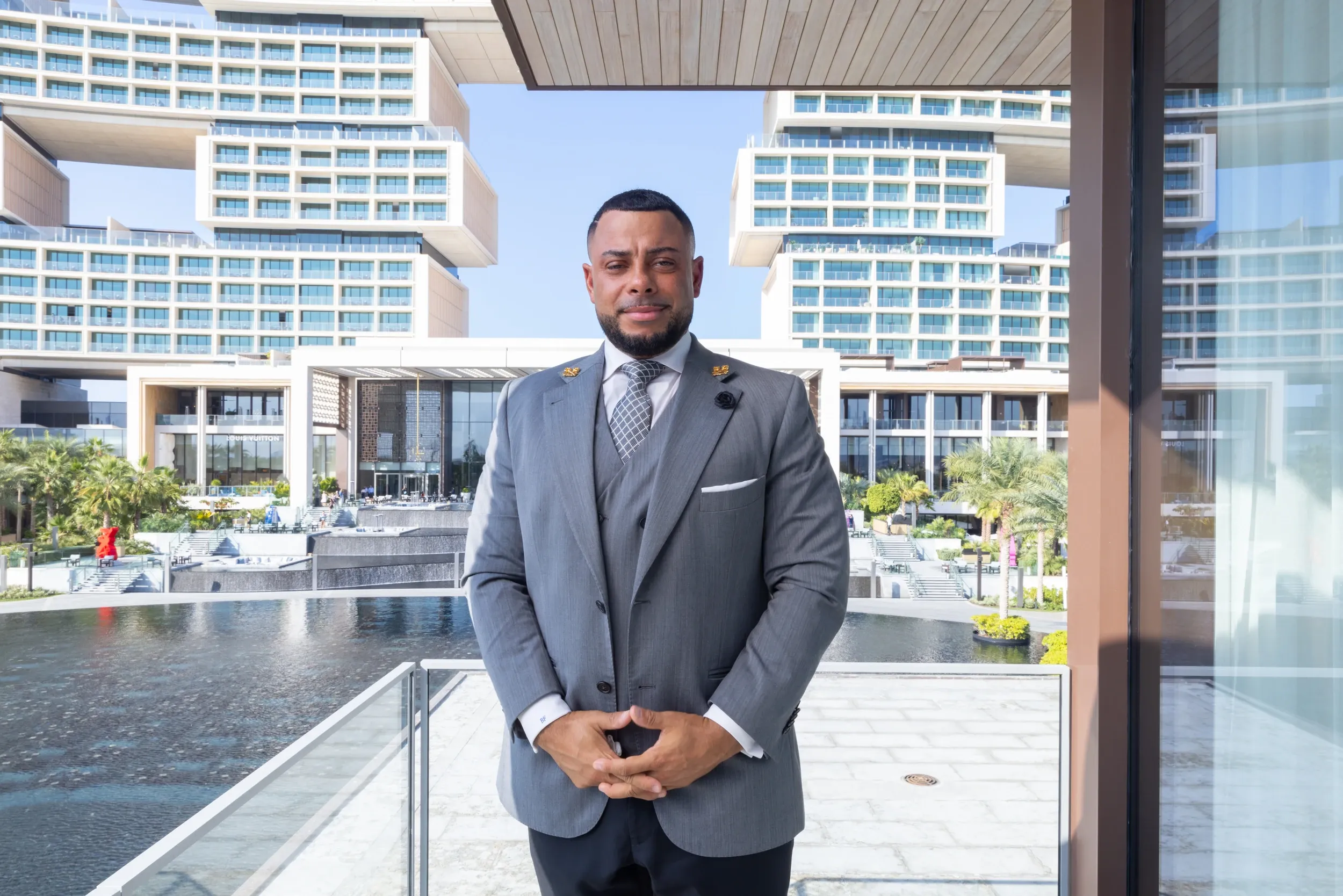 A man in a grey three-piece suit on a terrace overlooking a contemporary hotel.