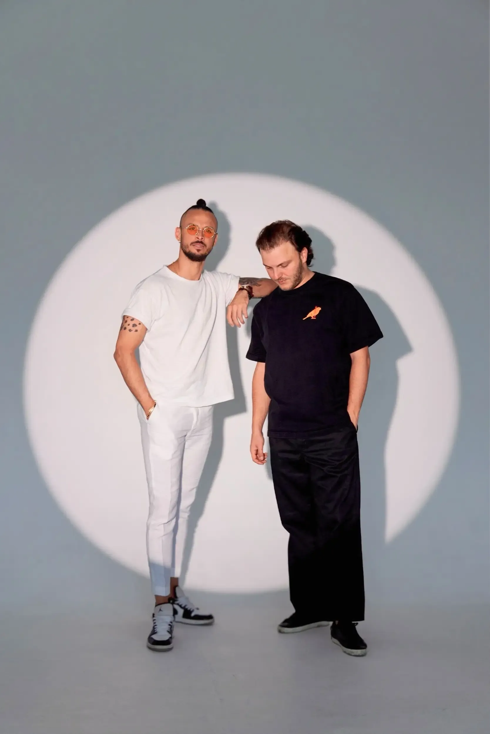 Studio duo portrait: one man in all white, one in black, under a circular spotlight