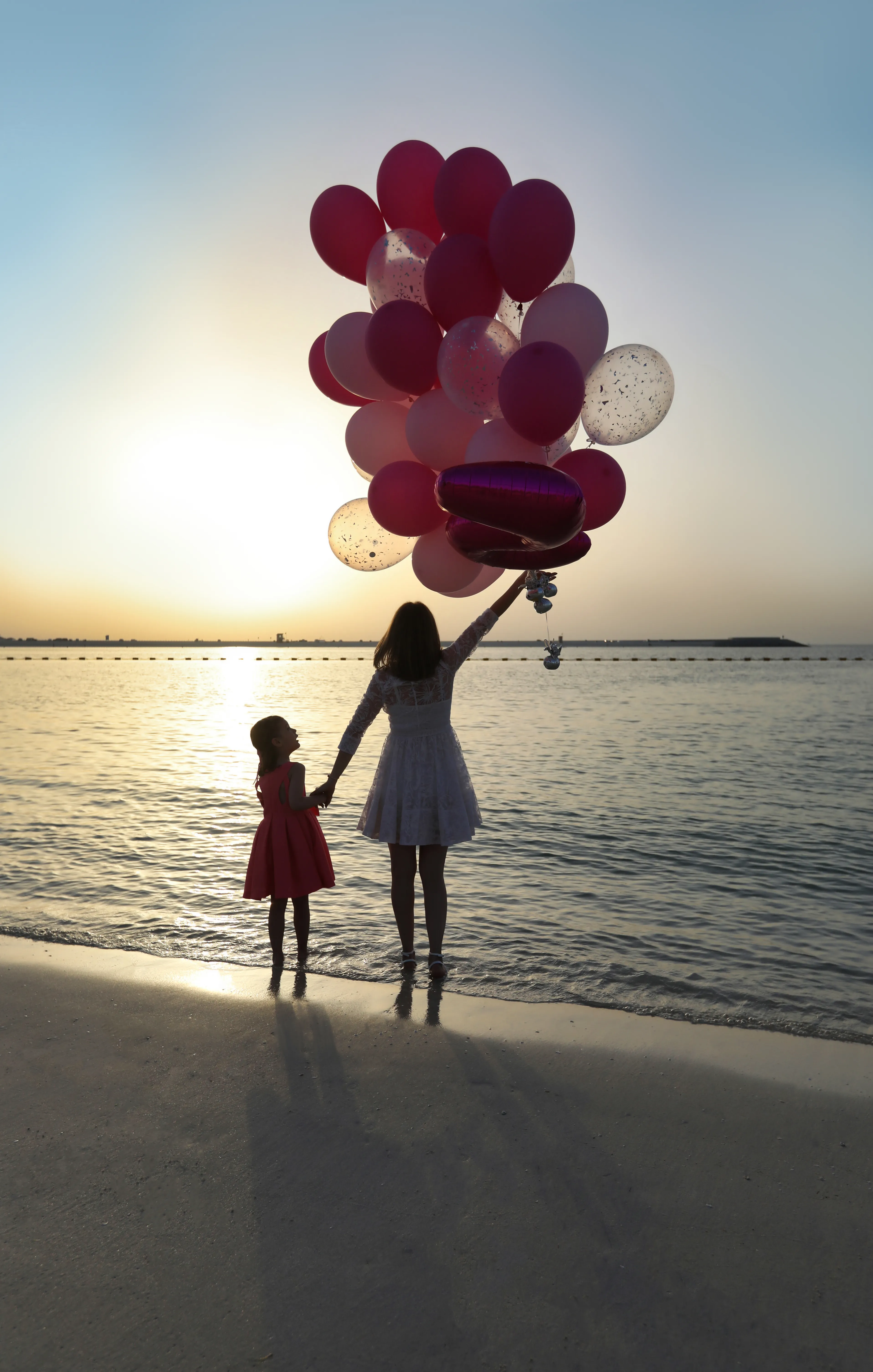 Mother and daughter with balloons at beach sunset, silhouettes