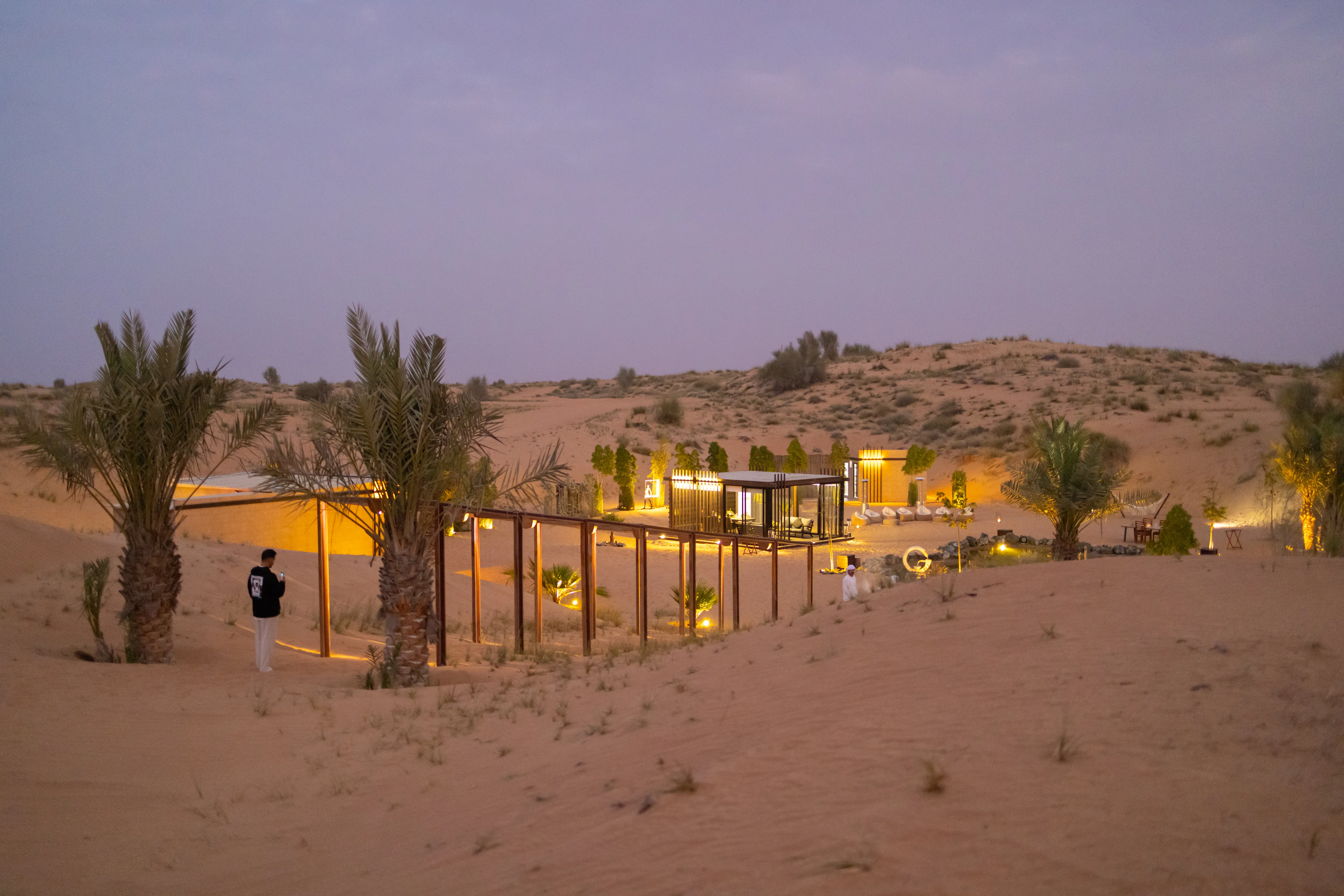 An illuminated desert venue among dunes and palms at dusk.