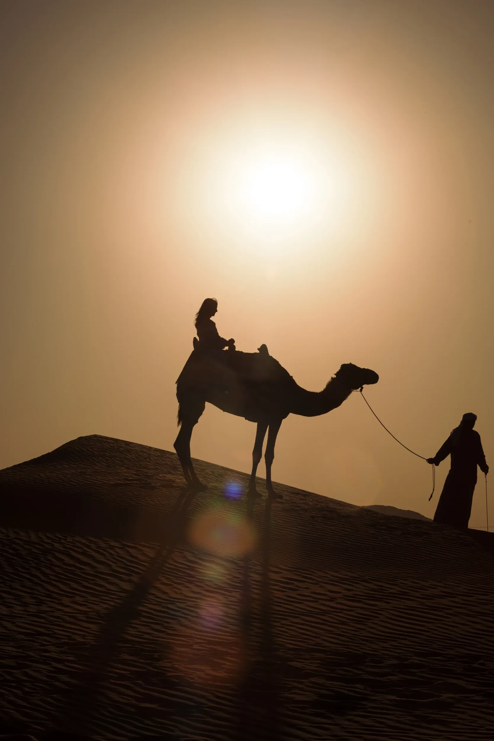 A camel, rider and handler silhouetted beneath a low sun on a dune ridge.