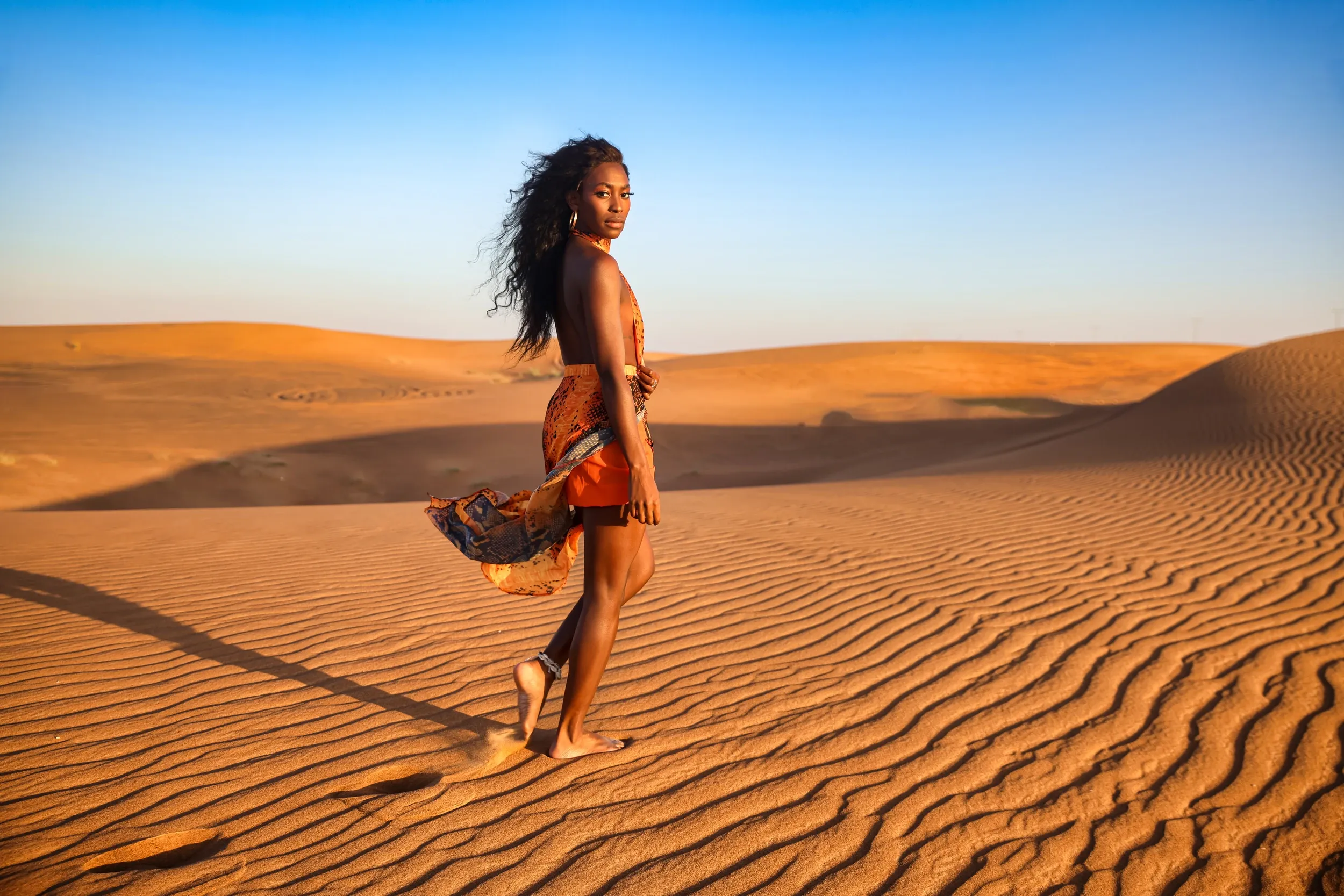 A woman in a patterned orange outfit looking back while walking across sunlit dunes.