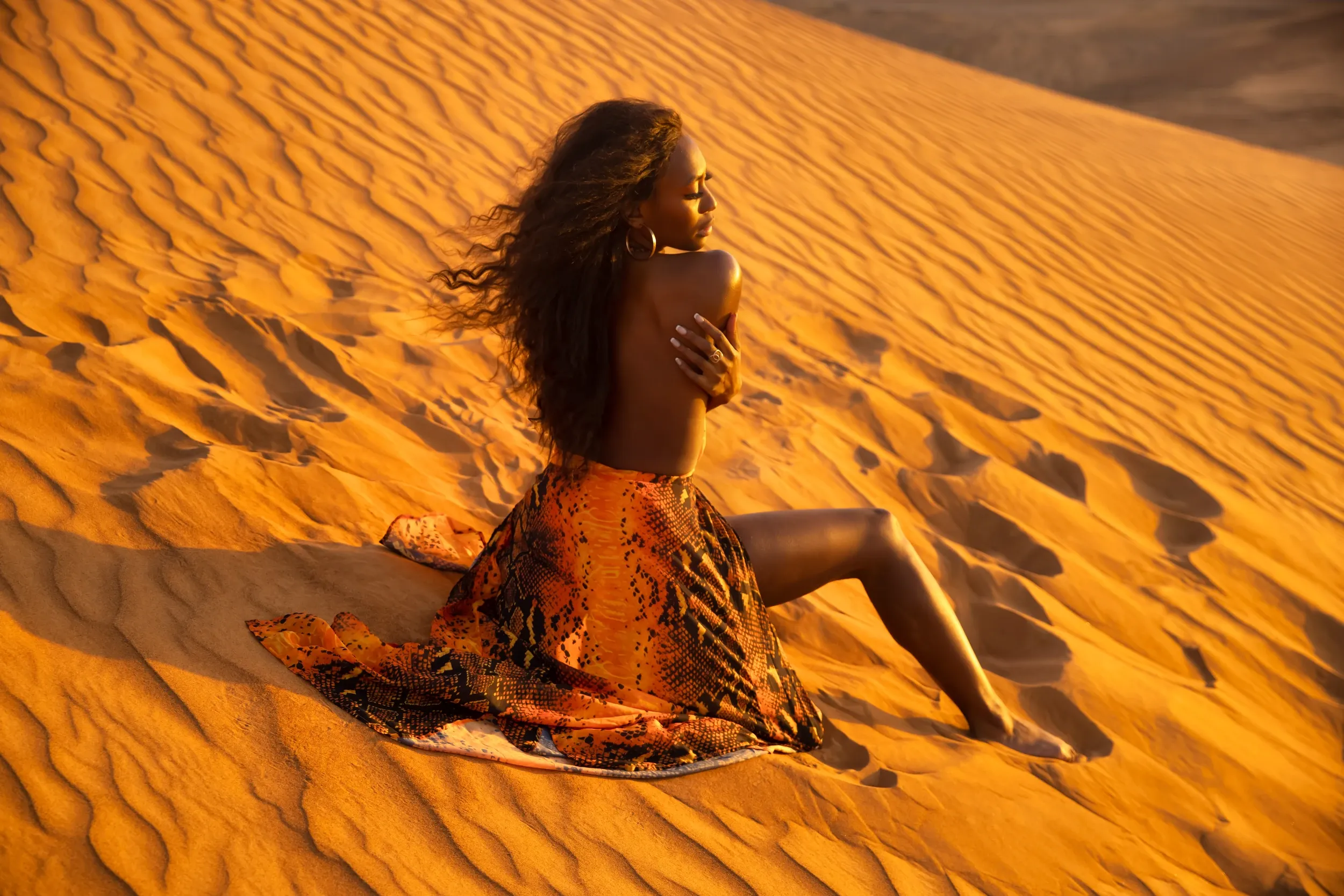 A woman seated on glowing orange sand with patterned fabric trailing behind her.
