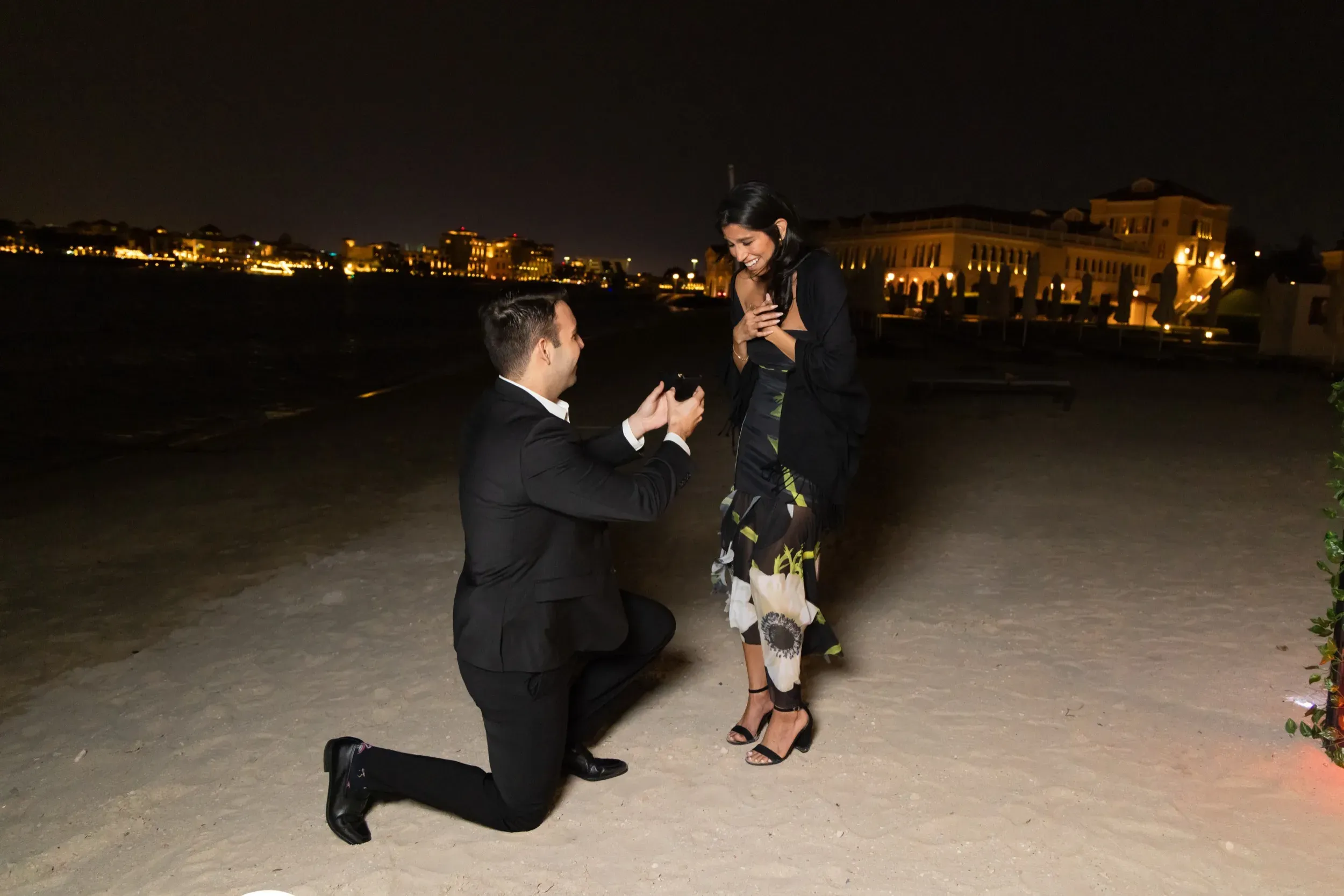 Night beach proposal: man on one knee with open ring box as she reacts with joy outside a lit villa