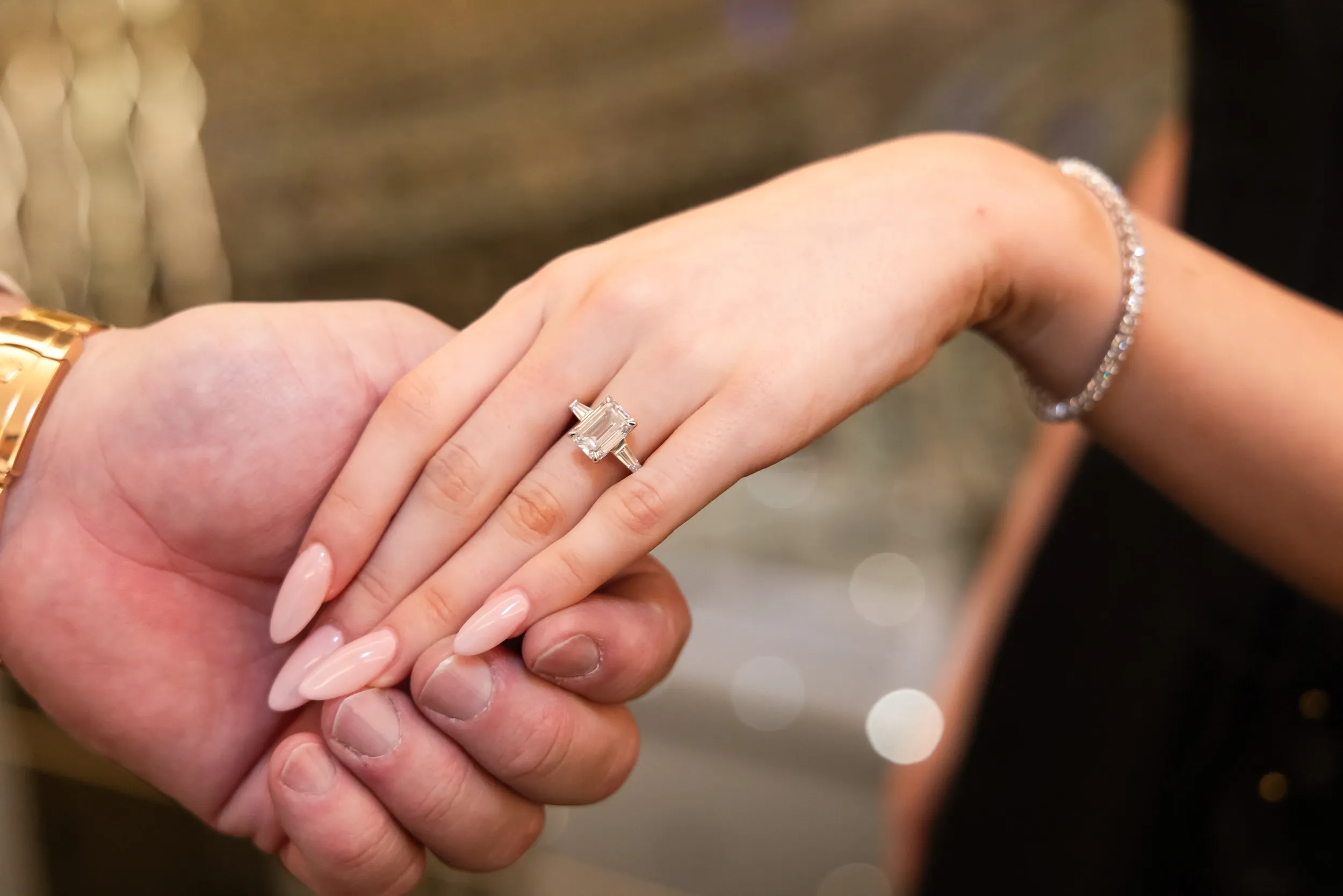 A close-up of joined hands displaying an engagement ring against a softly blurred warm background.