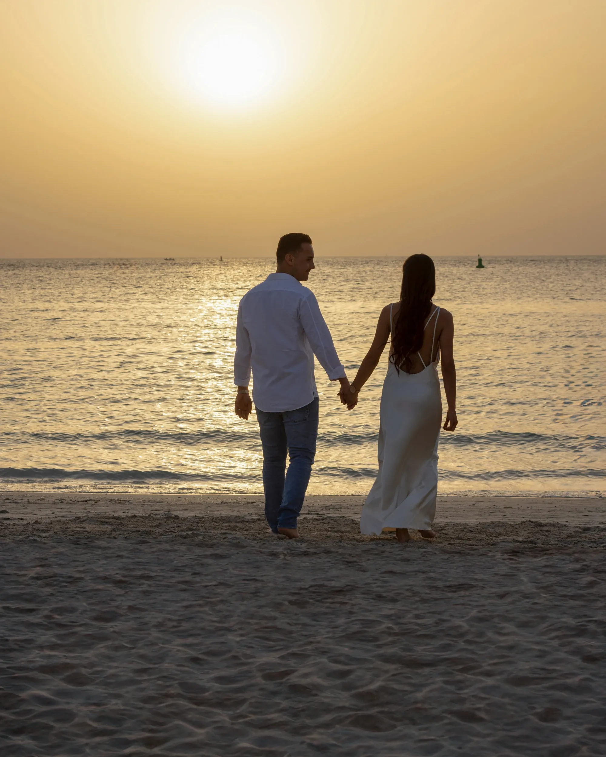 A couple holding hands while walking toward the sea beneath a low sun.