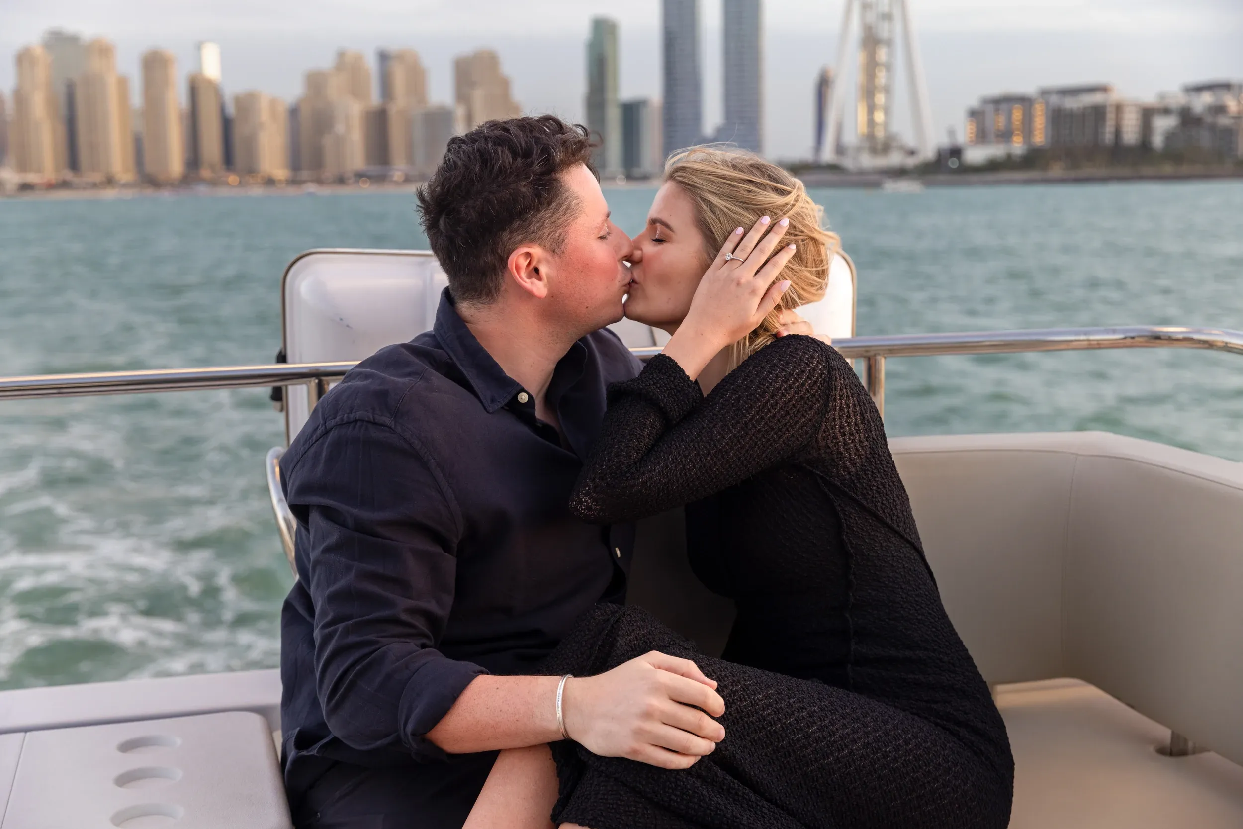 A couple kissing while seated on a yacht with waterfront towers behind.
