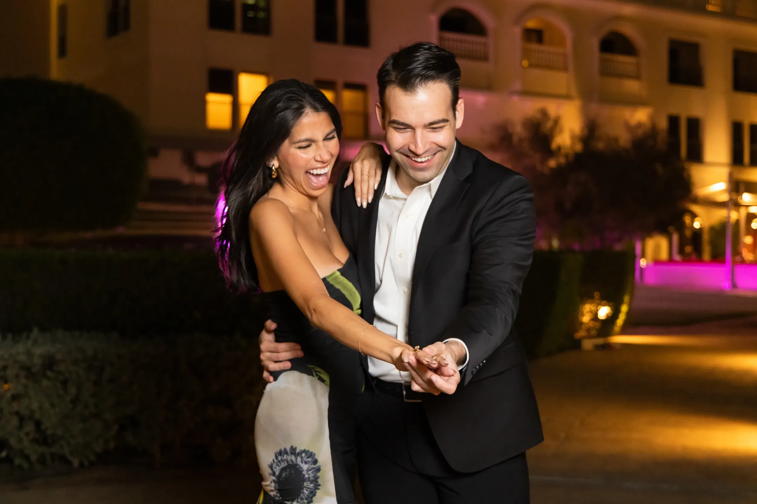 A couple laughing while looking at an engagement ring outside a lit building at night.
