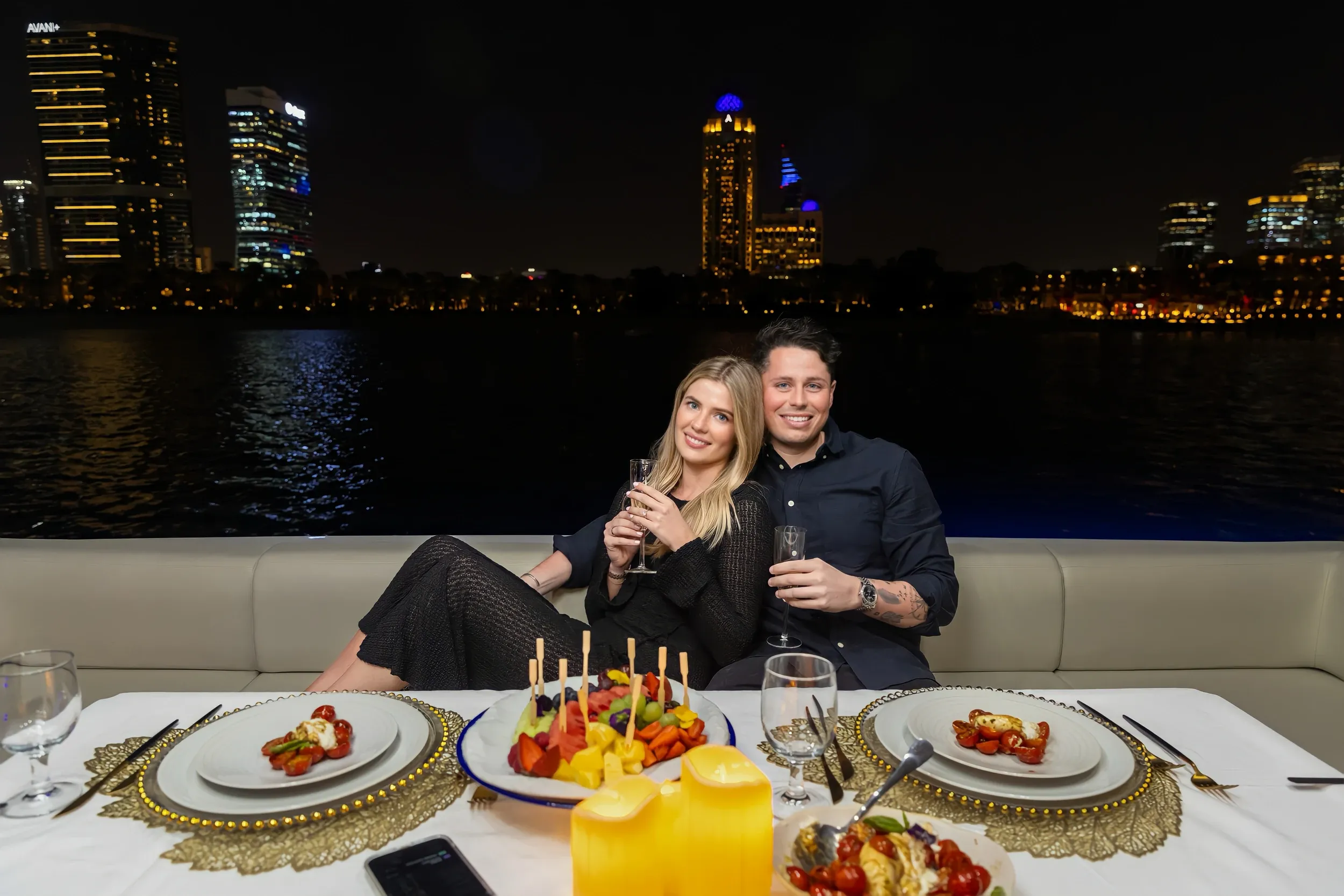 A couple seated behind a candlelit dinner table on a yacht at night.