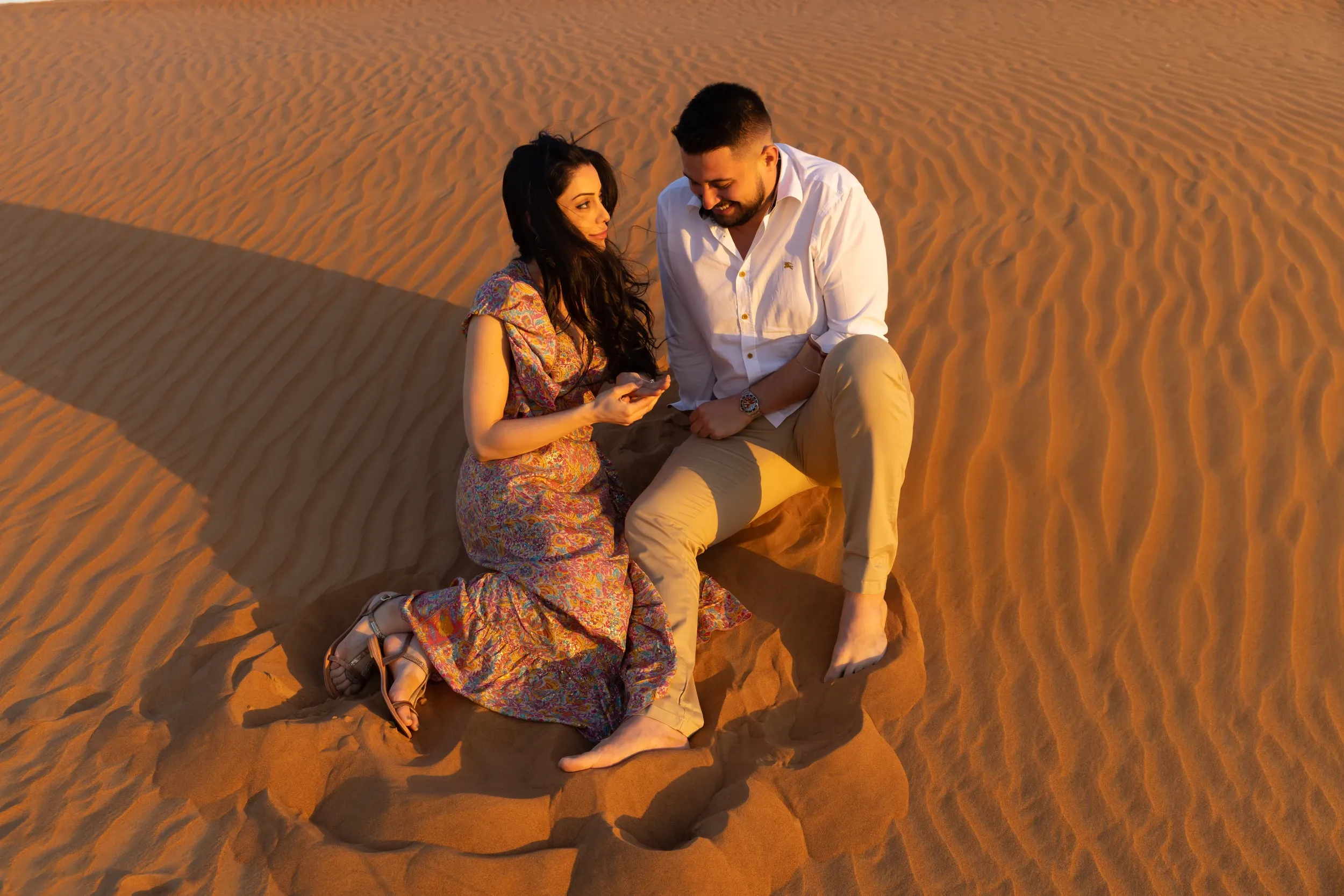 A couple seated and kneeling close together on rippled sand, looking down toward their hands.