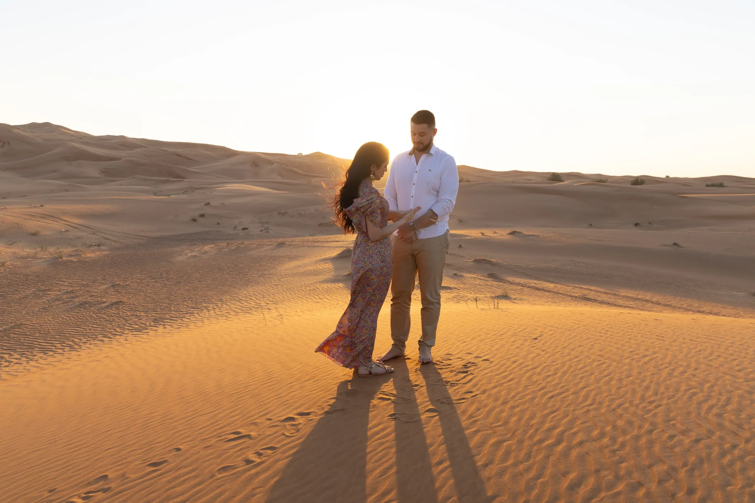 A couple standing together on sunlit dunes with long shadows stretching across the sand.
