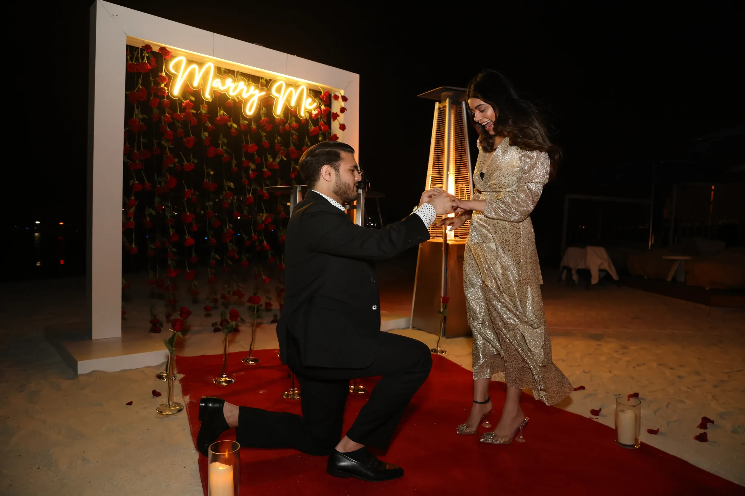 A man kneeling to propose beside a red carpet and illuminated Marry Me floral backdrop.