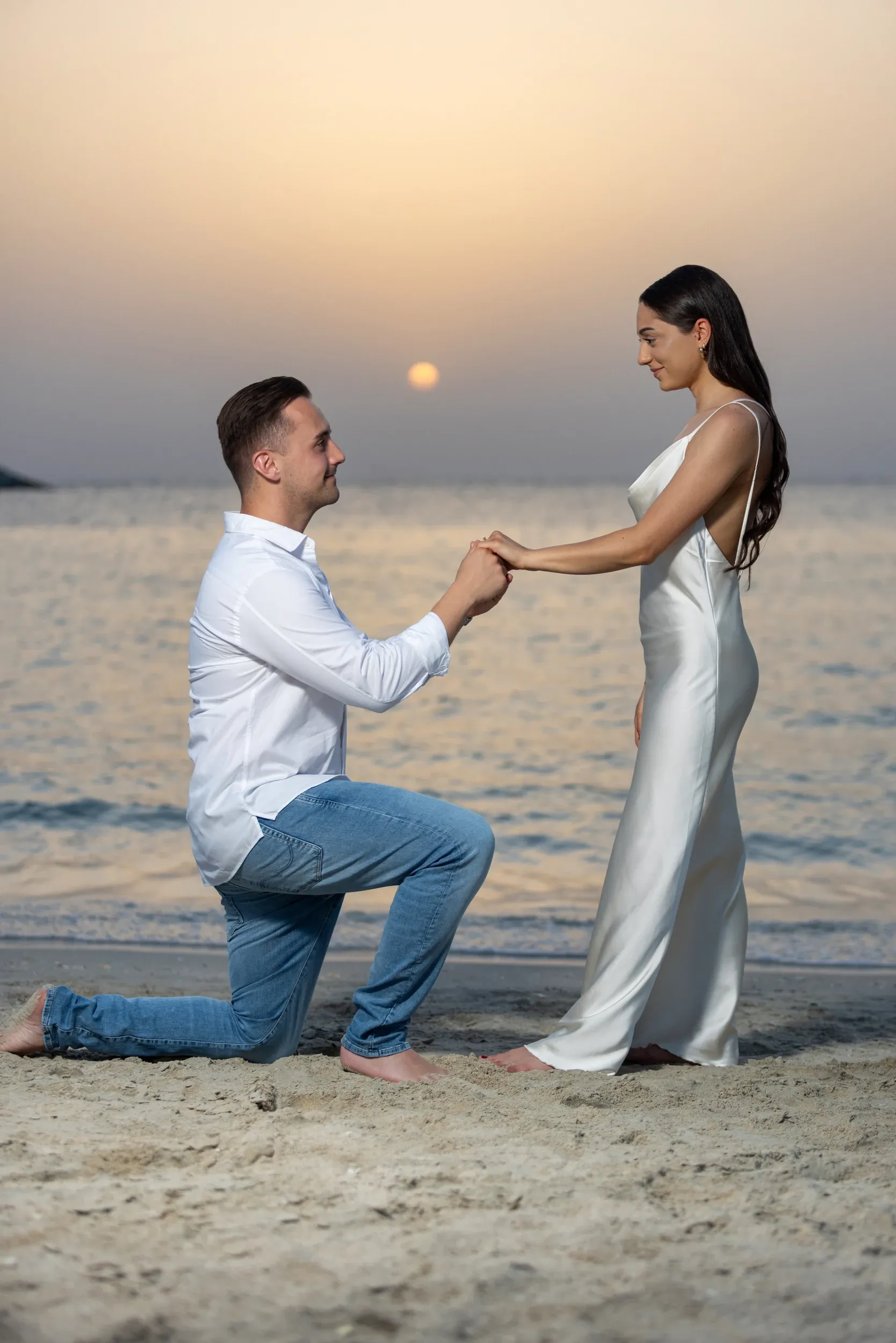 A man kneeling to propose to a woman on a beach at sunset.