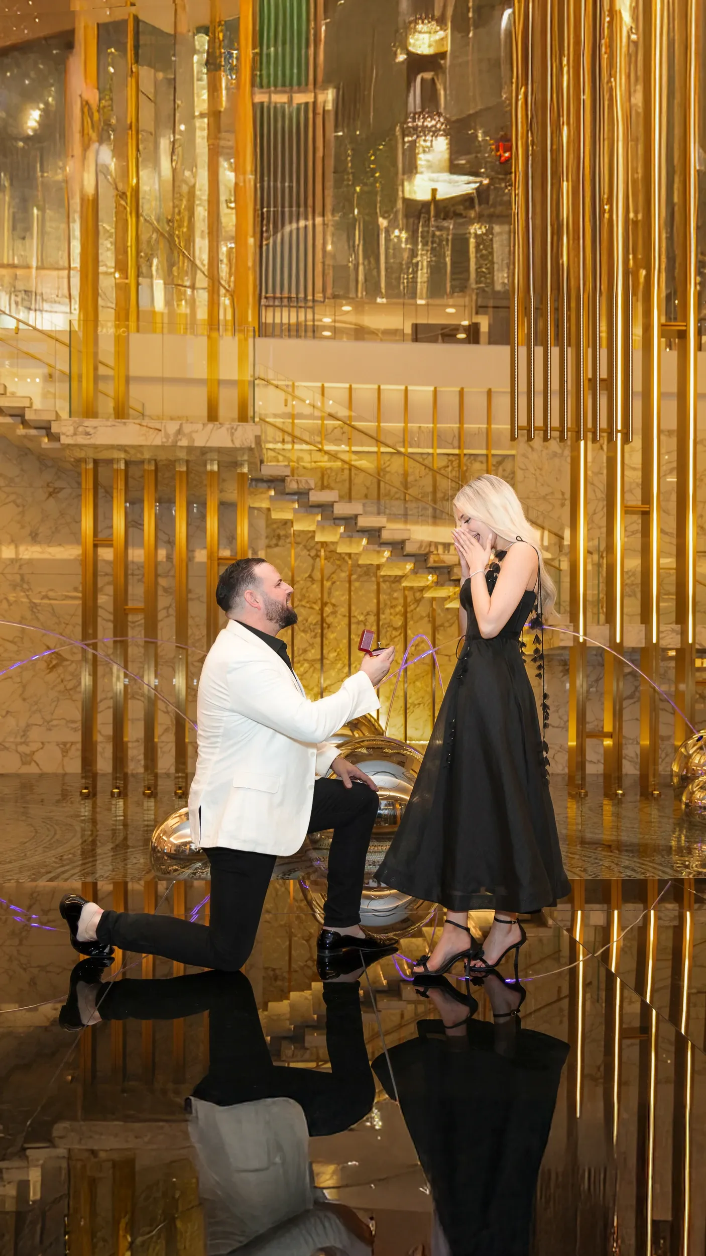 A man kneeling to propose to a woman in a black dress inside a reflective gold interior.