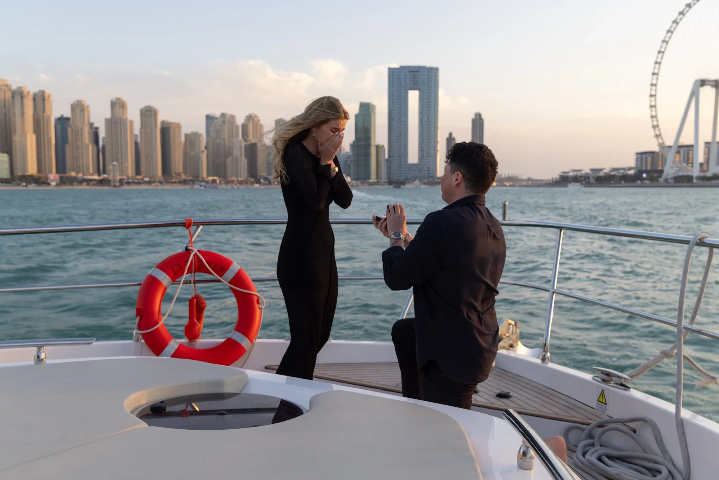 Yacht proposal on one knee with Dubai Marina skyline and Ain Dubai Ferris wheel behind at golden hour