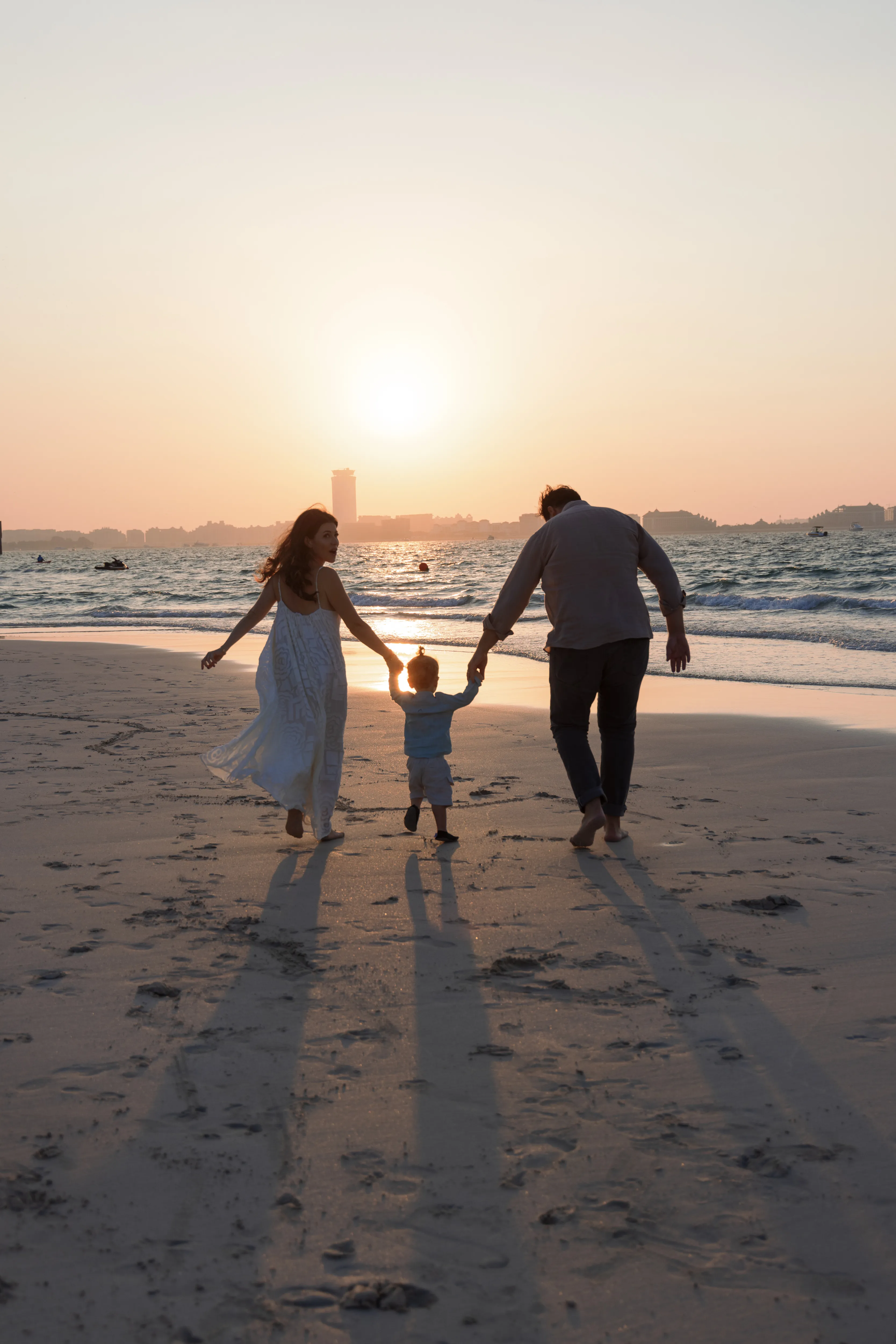 Two adults holding a small child's hands while walking toward the sea at sunset.