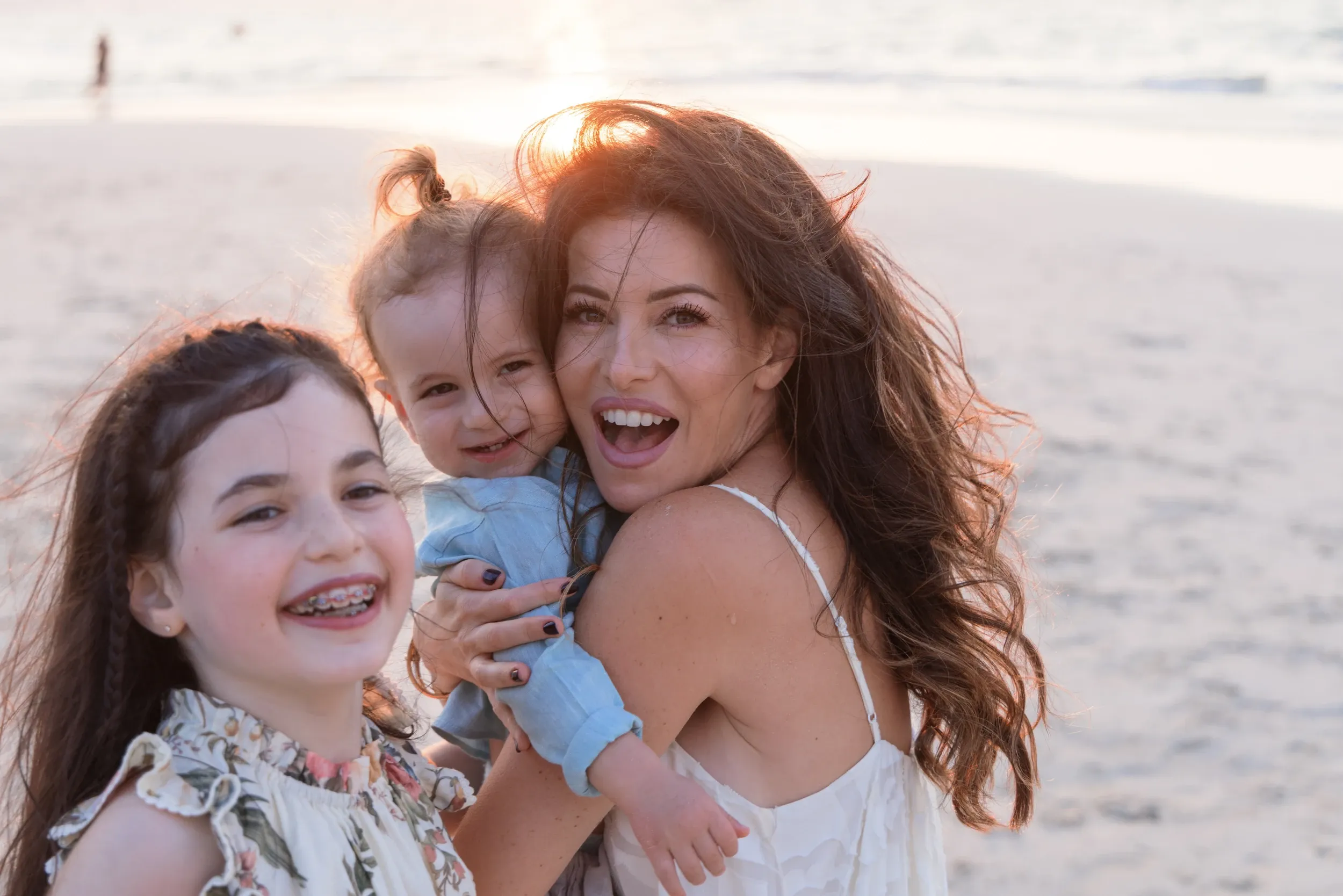 A woman and two children laughing together on a beach in warm backlight.