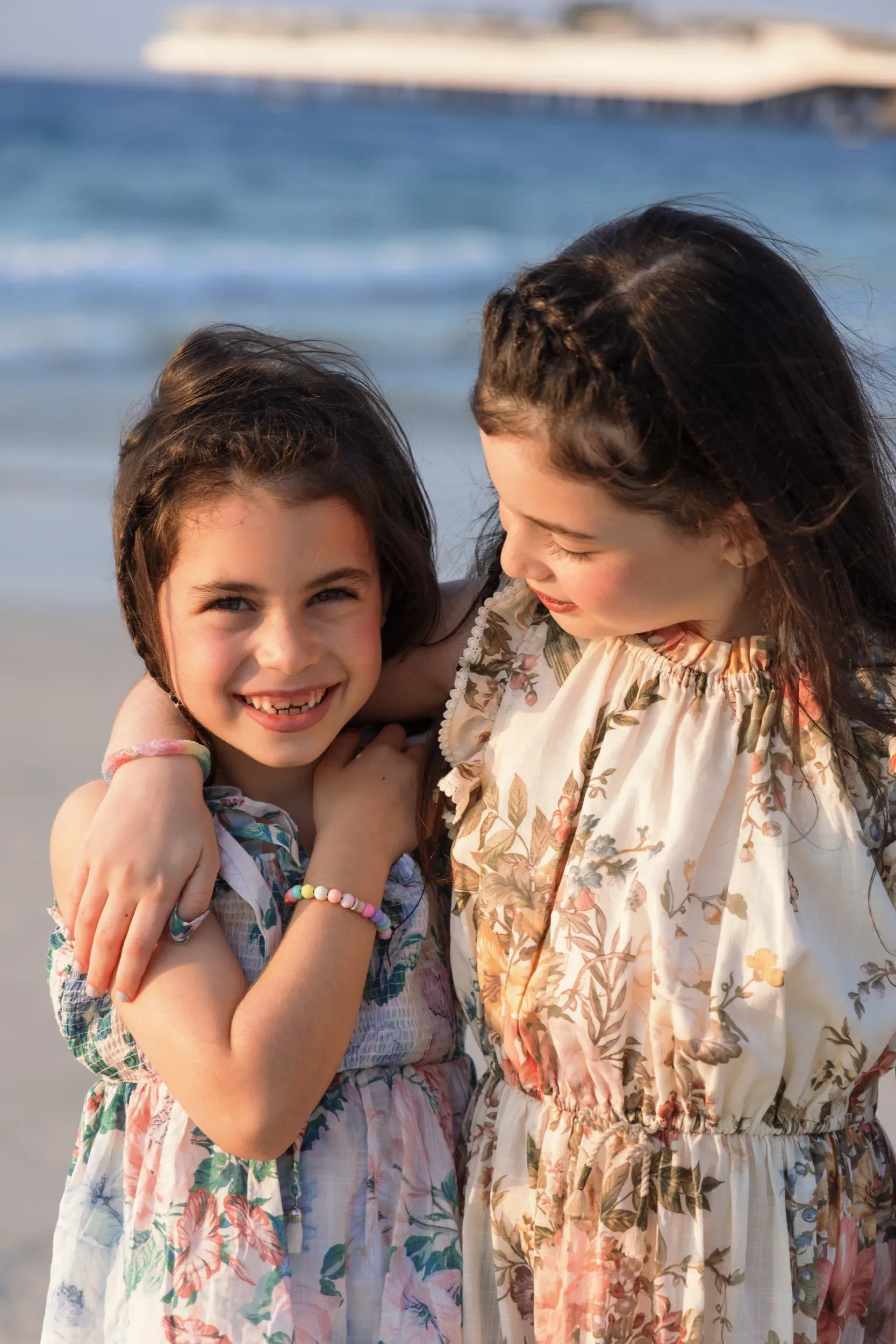 Two sisters in floral dresses embracing on the beach at golden hour