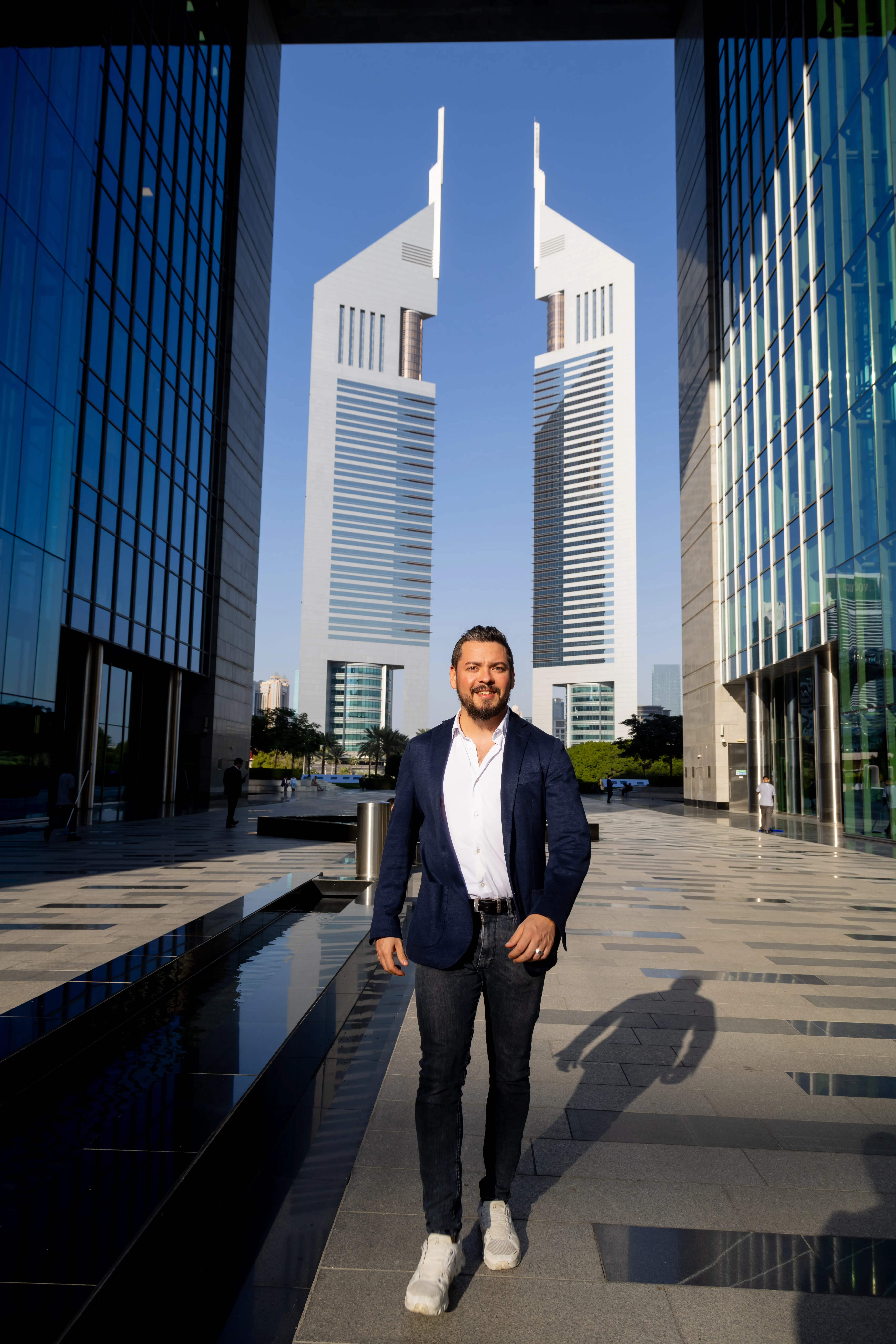 A man in a navy jacket framed by a tall gateway and glass buildings.