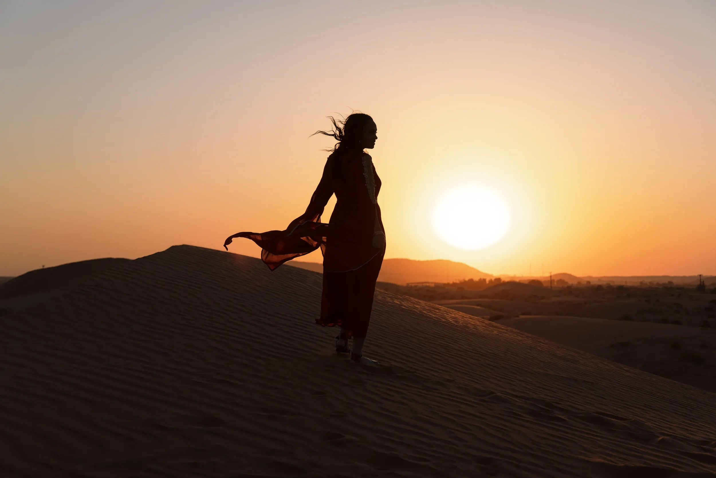 A woman silhouetted on a dune with loose fabric blowing beside the setting sun.