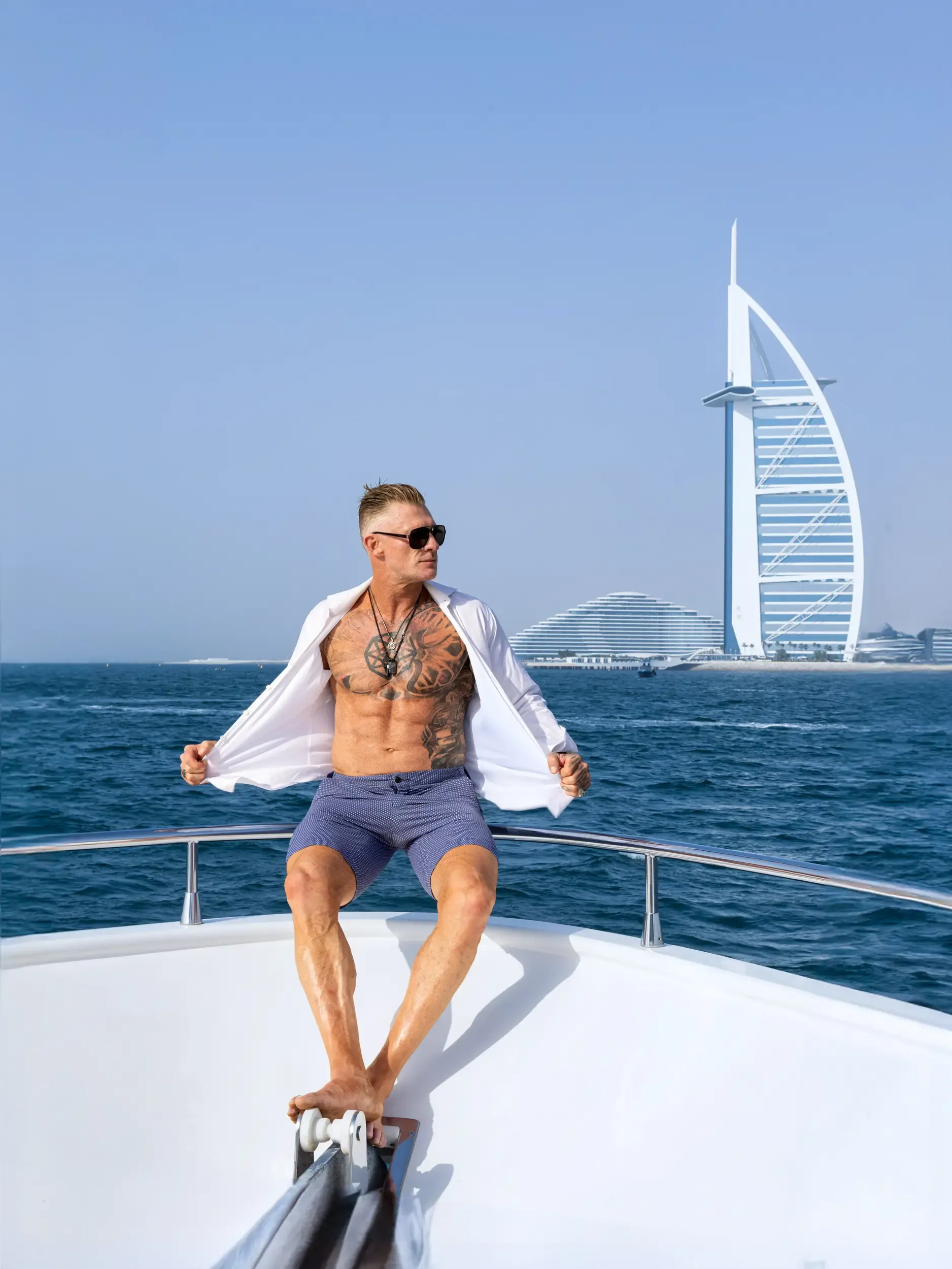 Shirtless man in open white shirt posing on a yacht bow with Burj Al Arab behind
