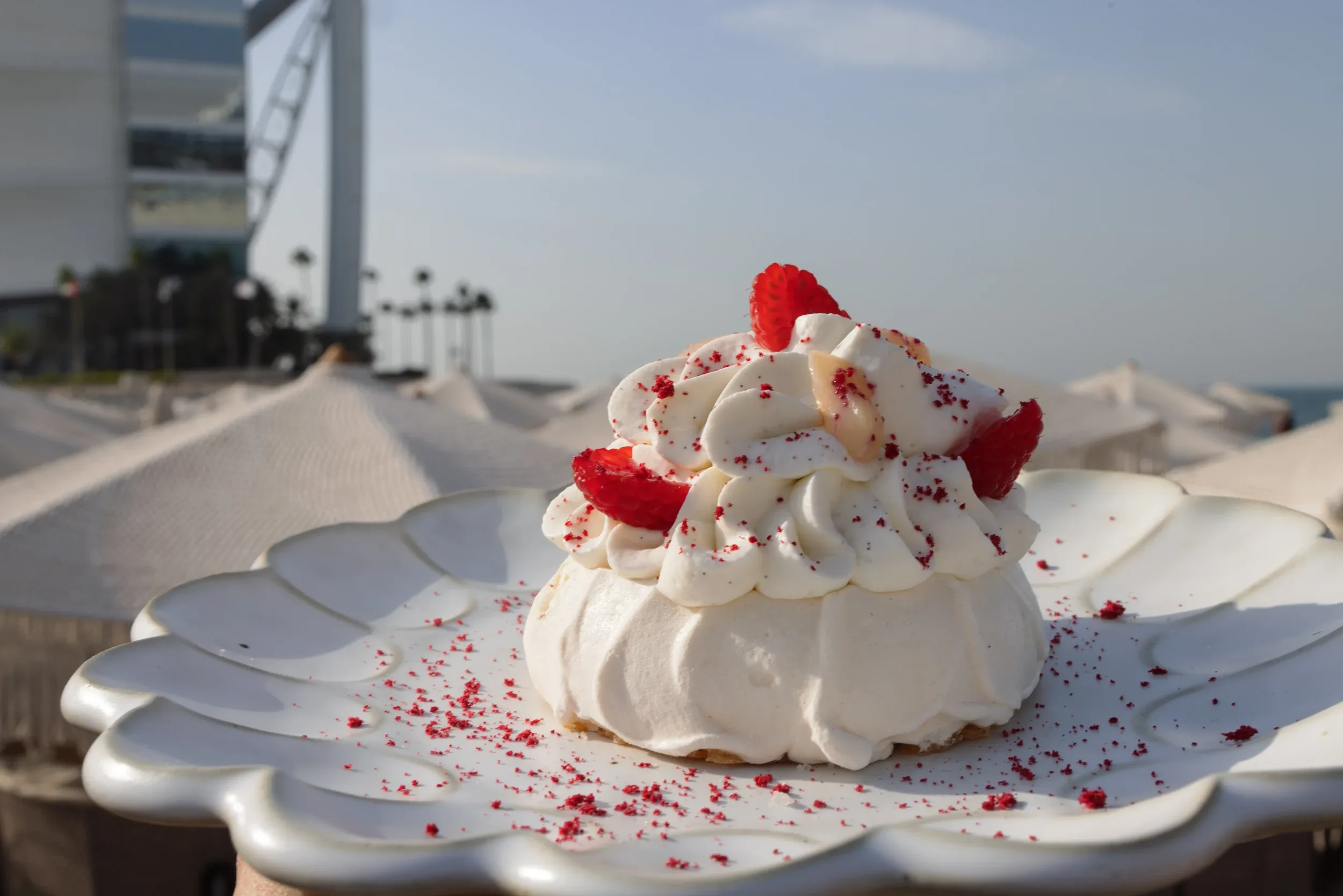 A white dessert with red berries on a sculpted plate, with the Burj Al Arab softly visible behind.