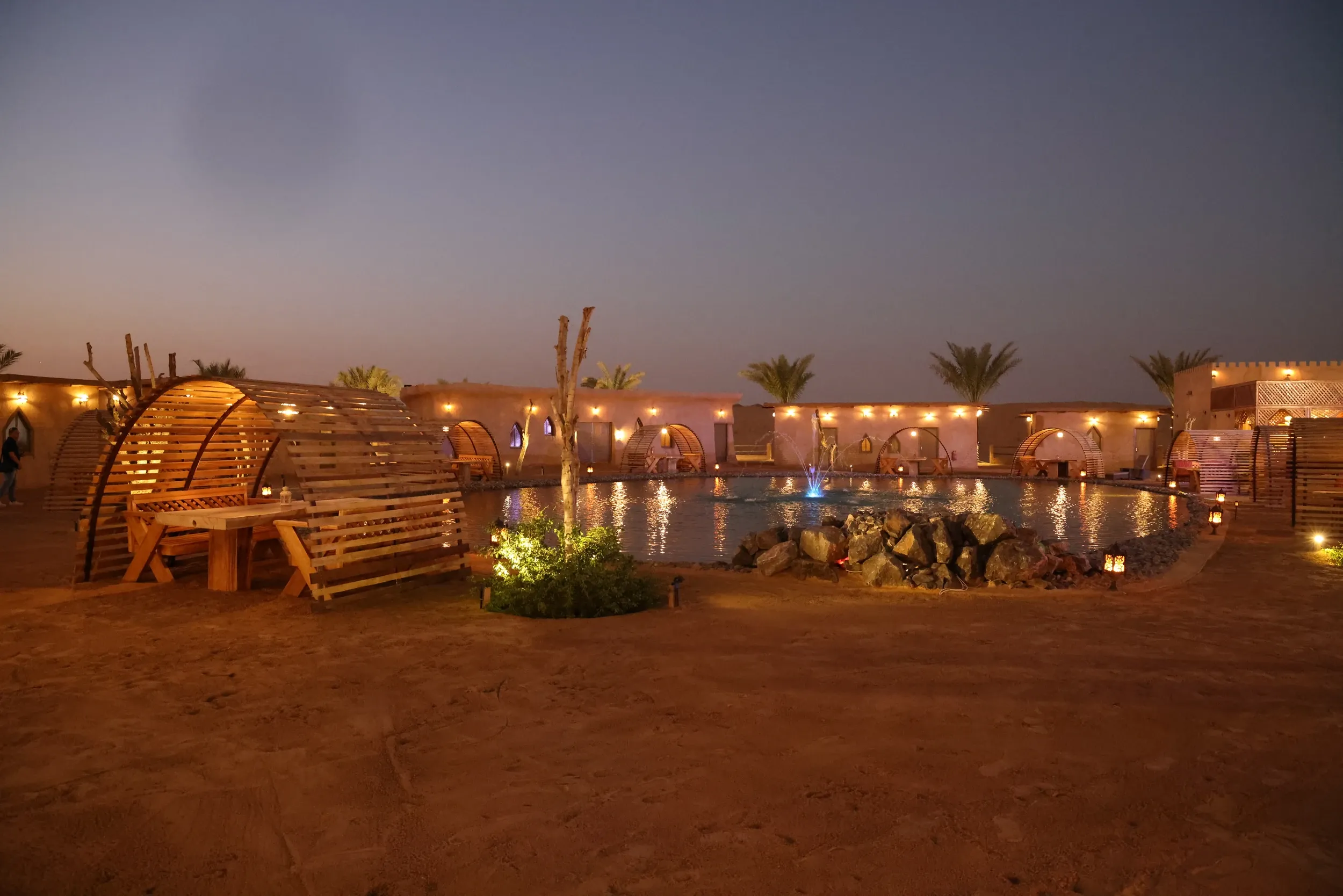 Warmly lit cabanas and seating around a desert-style pool at dusk.