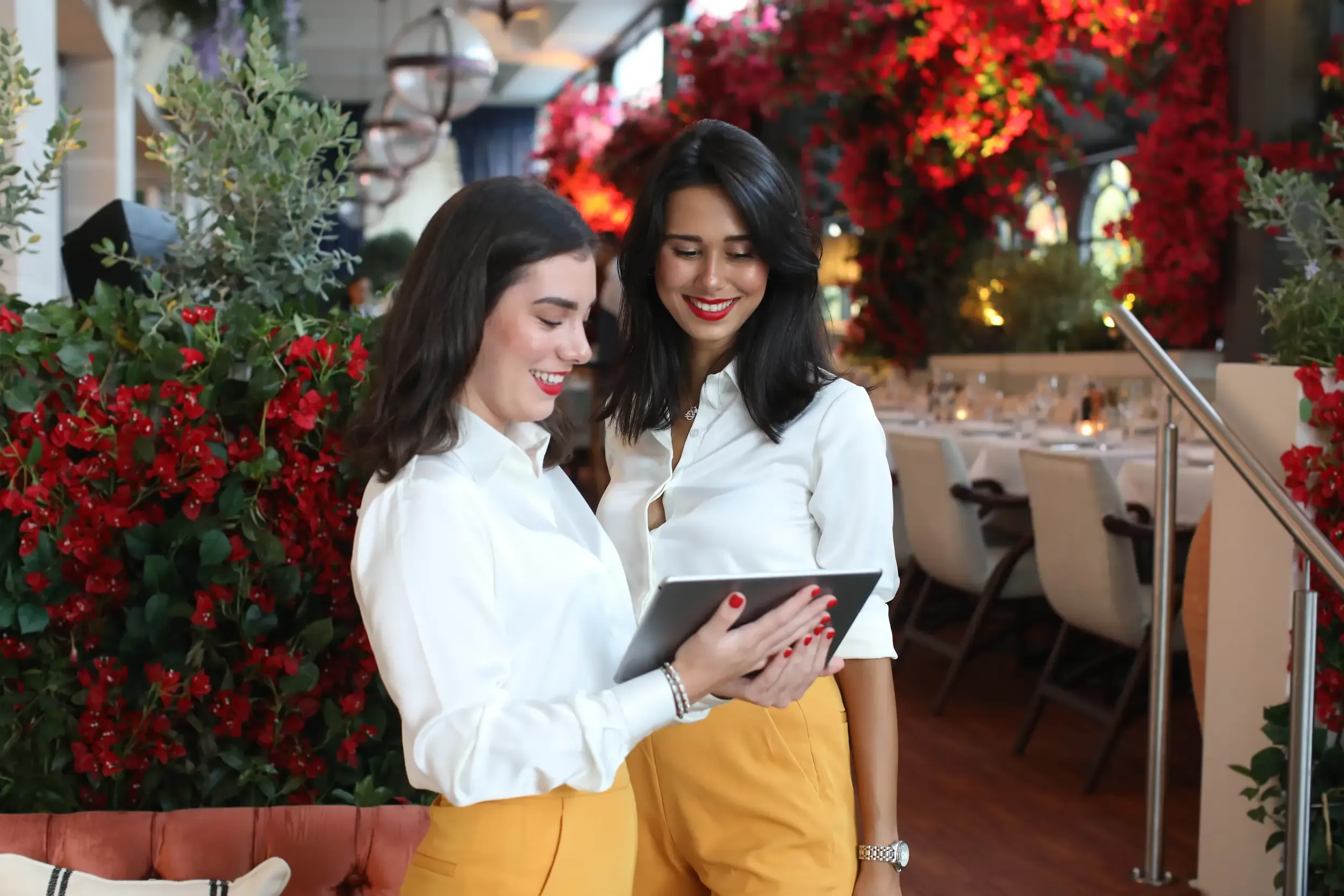 Two hostesses in white shirts and mustard trousers reviewing a tablet amid dining tables and red flowers