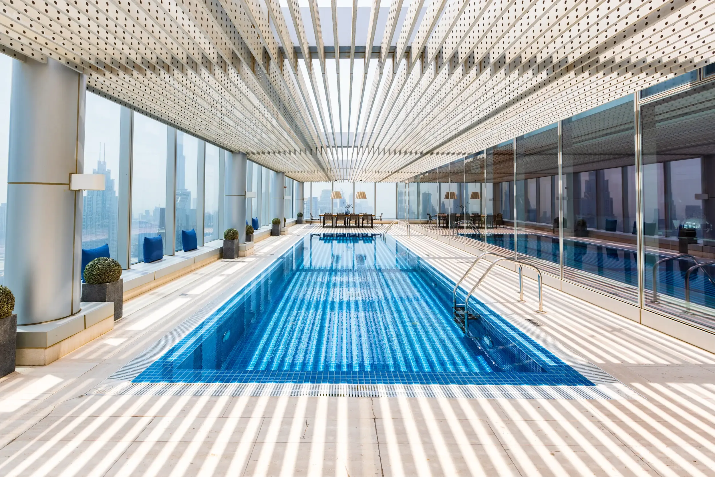 Long Rolex Tower lap pool between mirrored wall and glass columns with blue cushions and city view