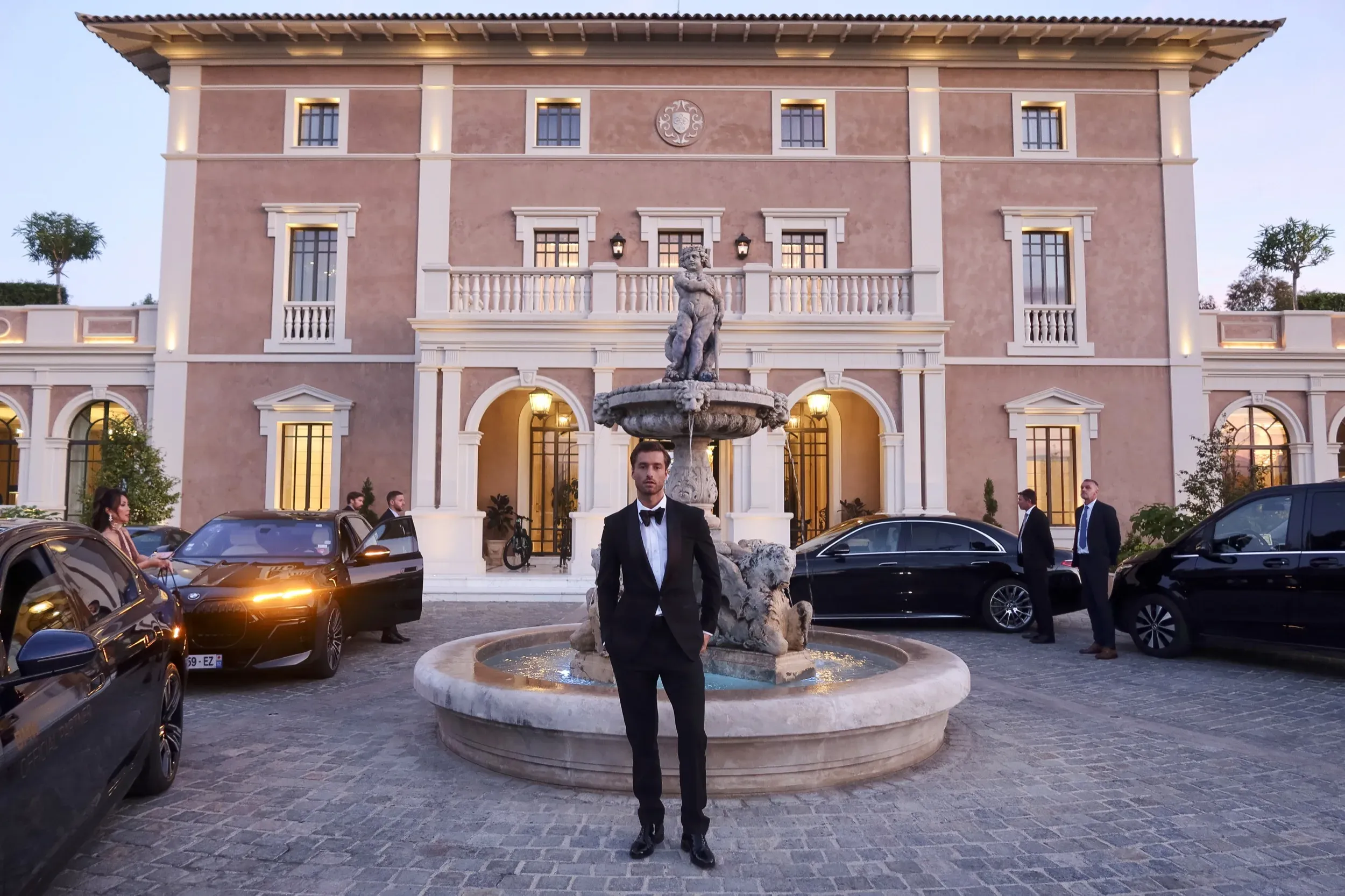 Man in a black tuxedo standing before a lit Mediterranean villa courtyard with fountain and luxury cars at dusk