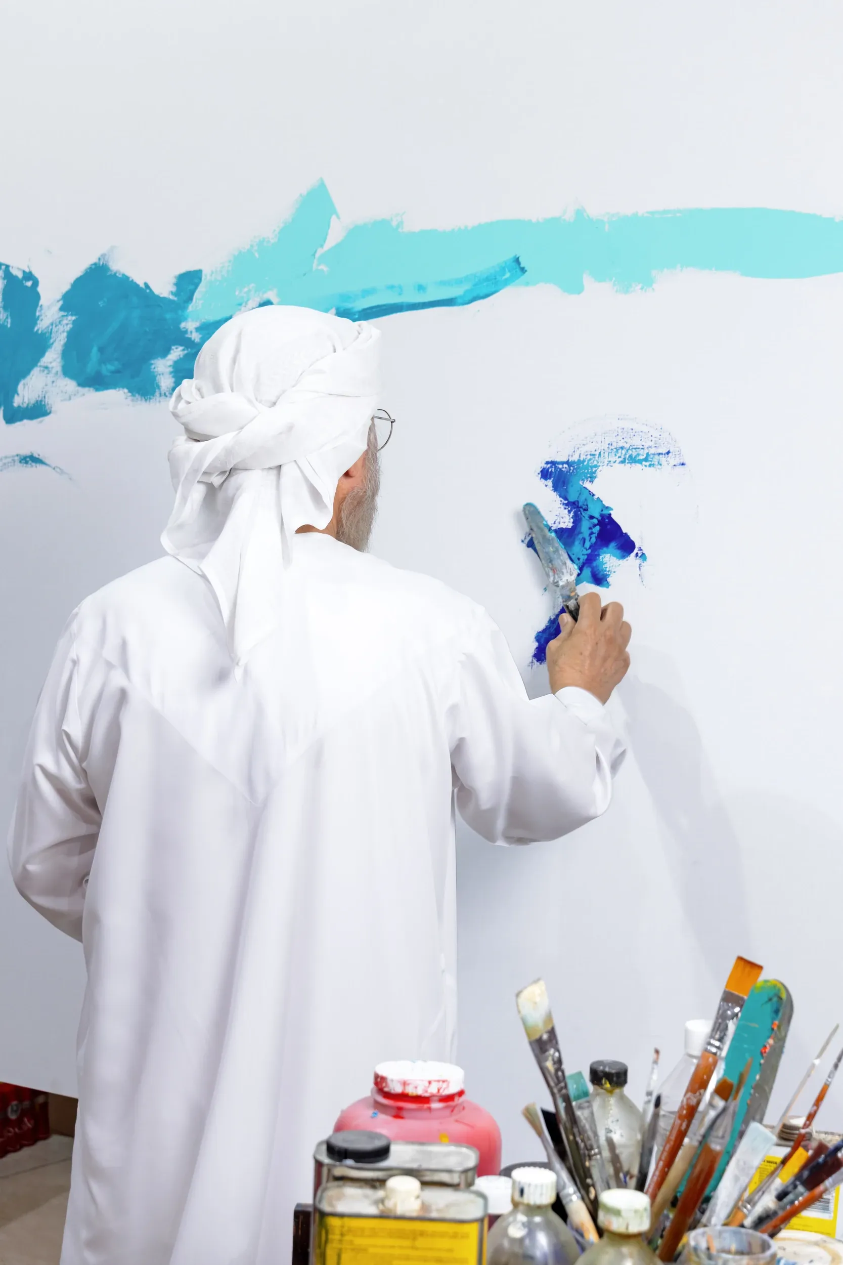 An artist in white applying blue paint to a large canvas, with tools in the foreground.