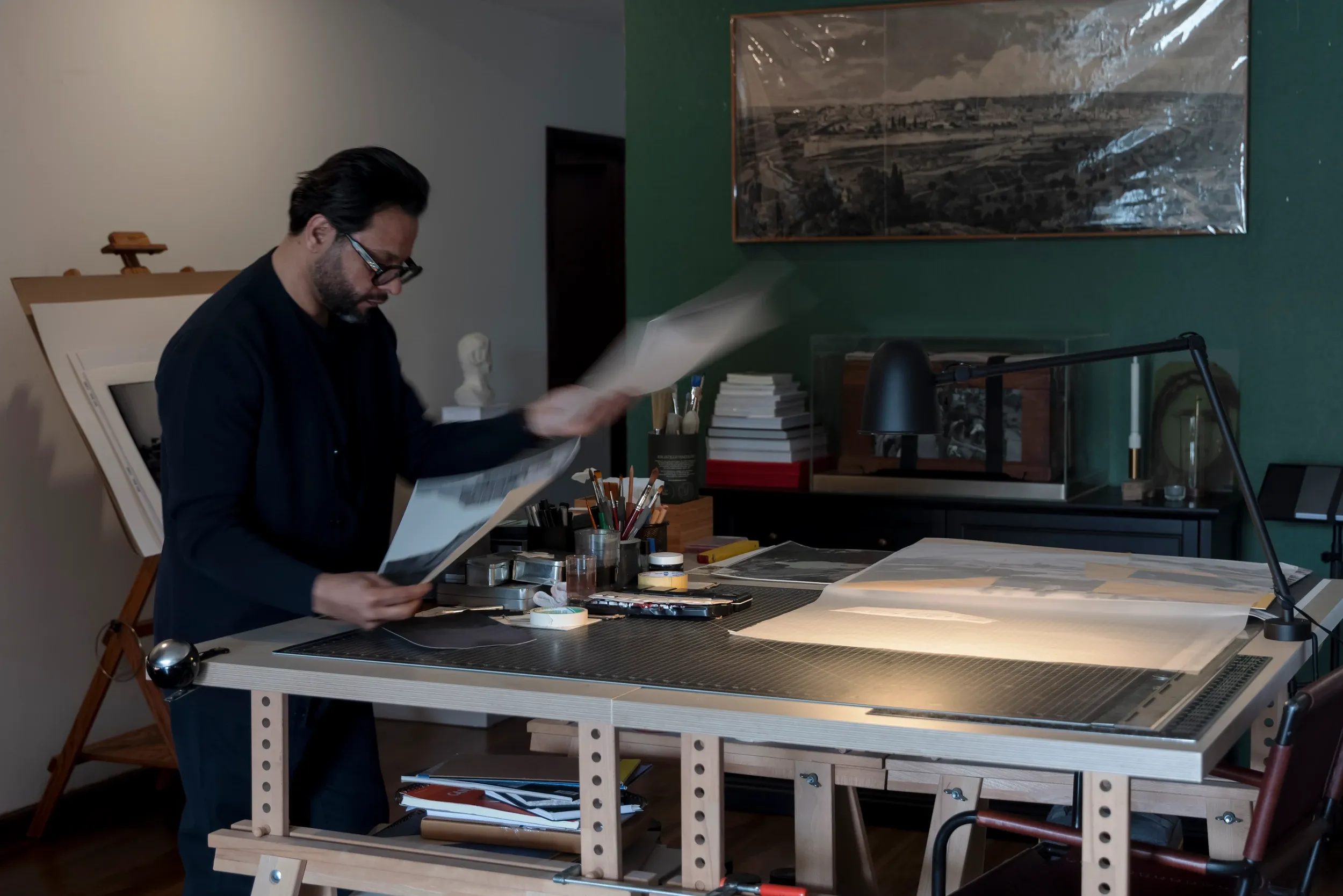 A man handling large sheets at a drawing table in a softly lit studio.