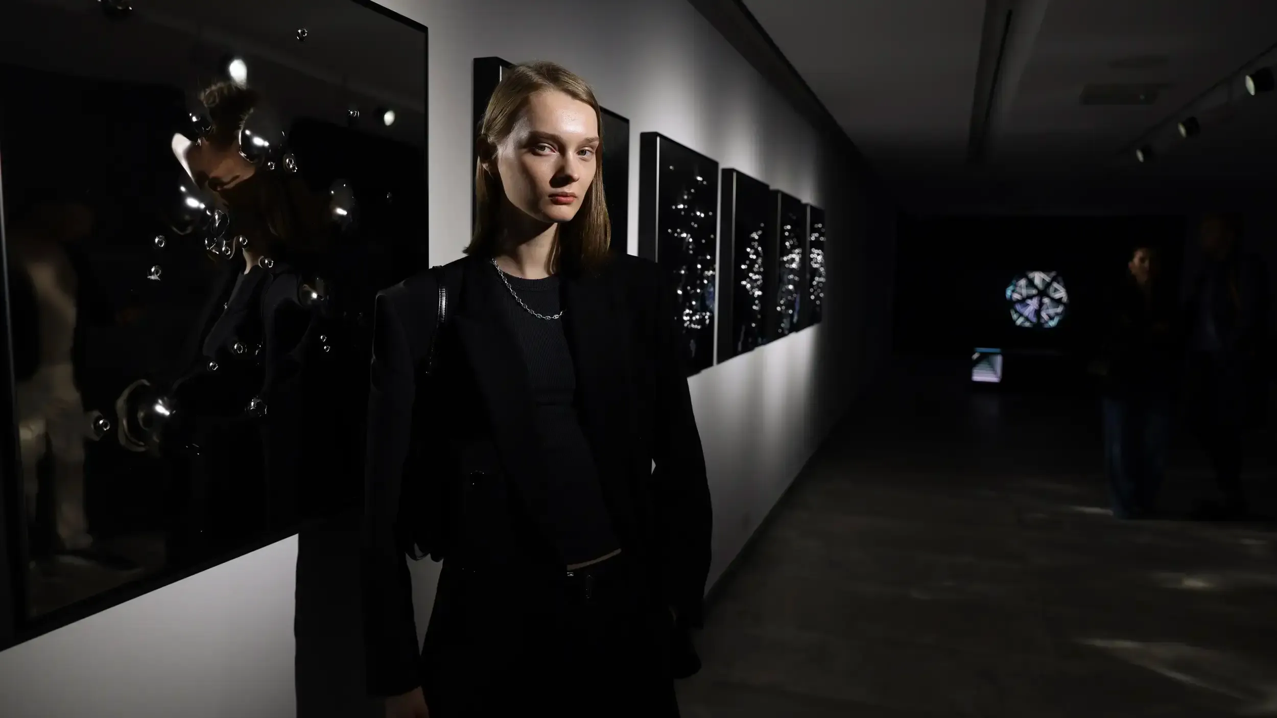 Woman in a black blazer standing before dark reflective artworks in a dim contemporary gallery