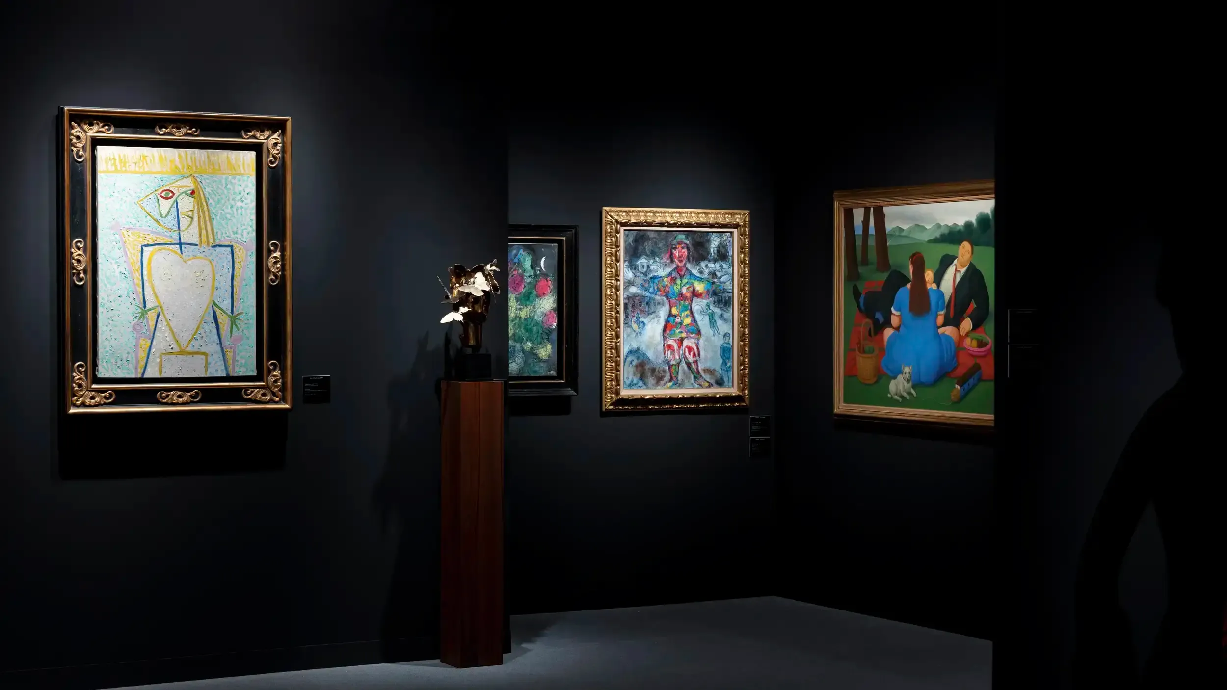 Dim gallery hang with Cubist and Botero-style figurative paintings and a gold metal sculpture on a pedestal