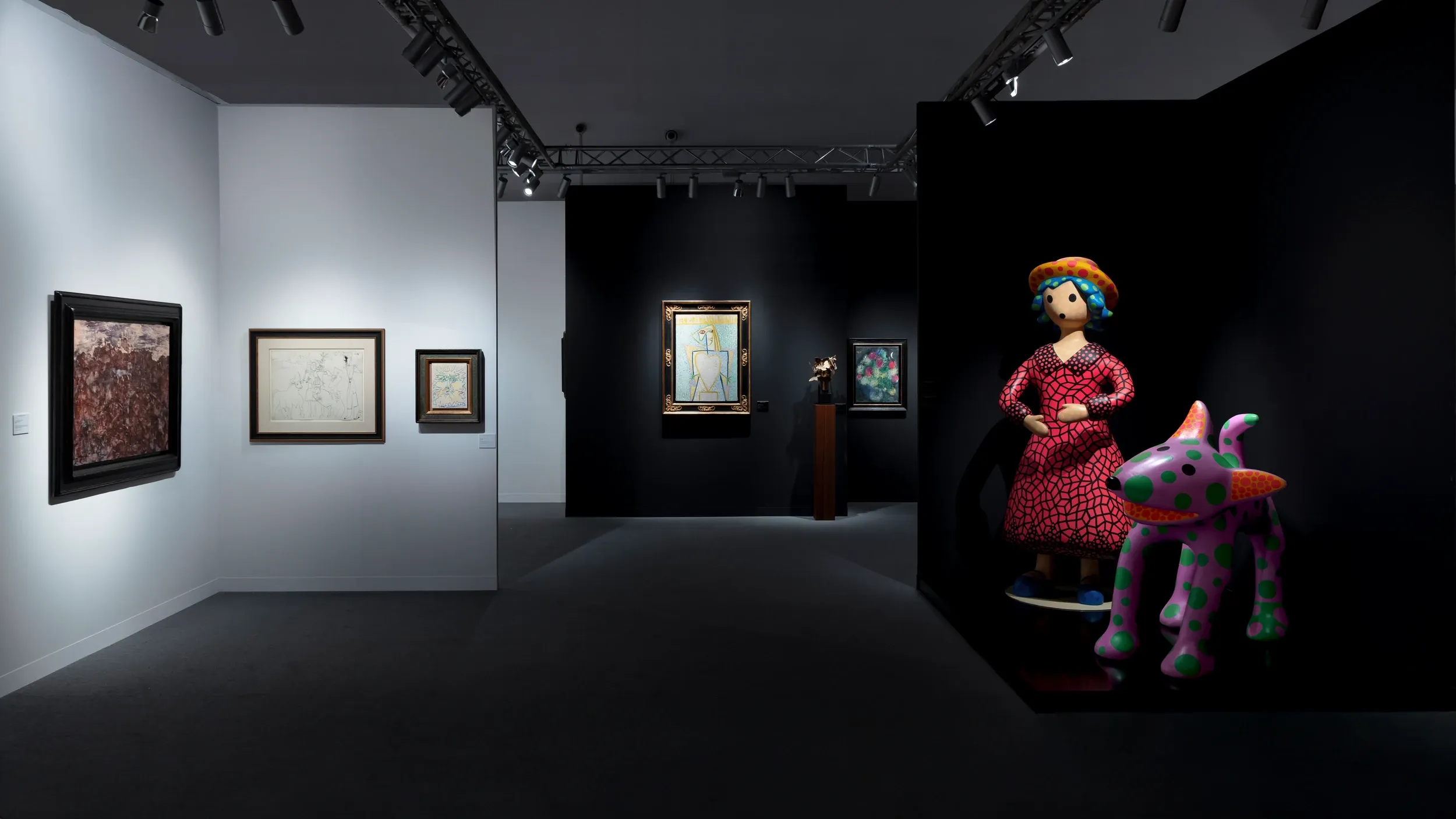 A wide exhibition installation with framed paintings and colourful sculptures under spotlights.